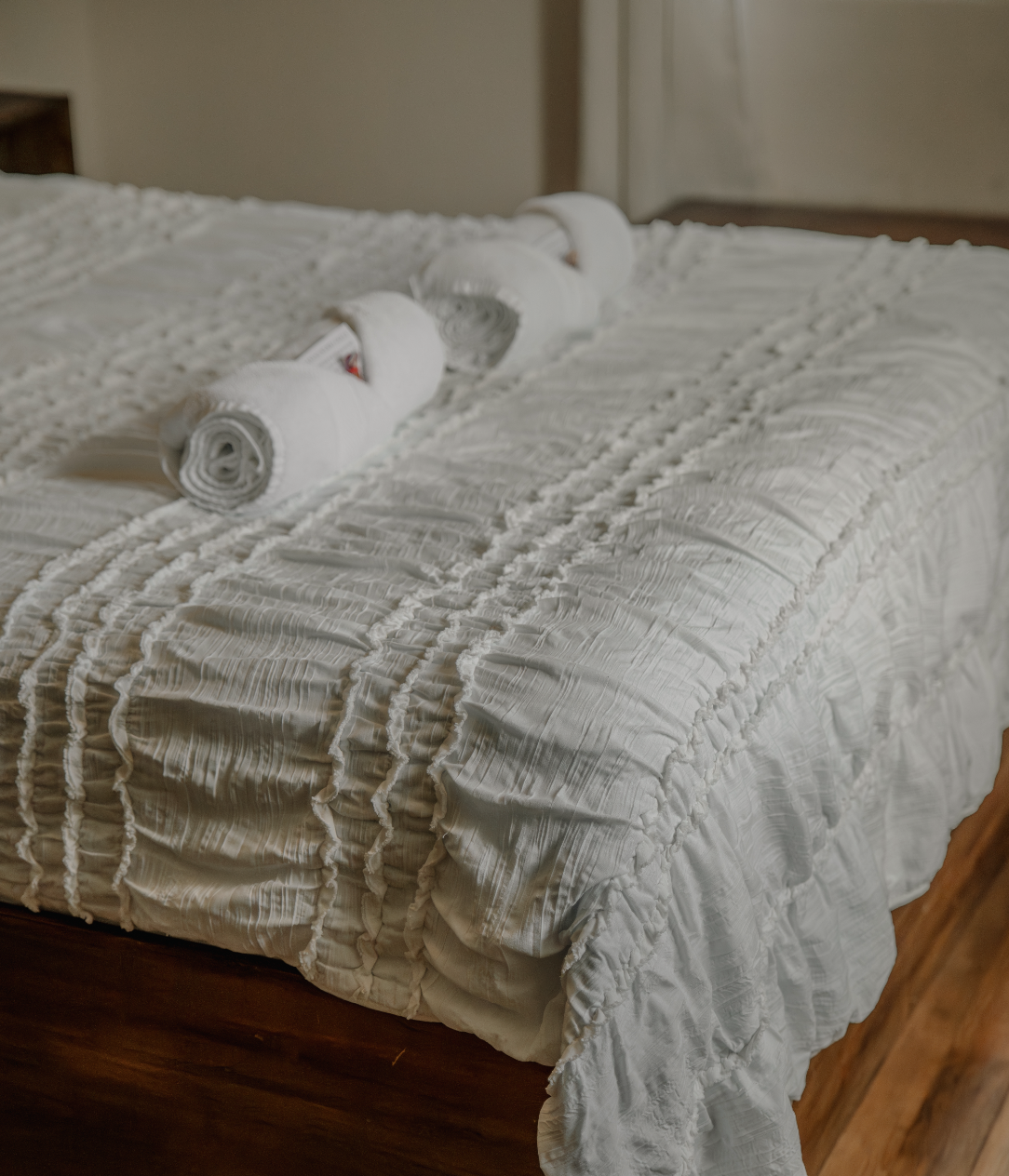 A neatly made bed with a white, textured bedspread and three rolled white towels placed across the top.
