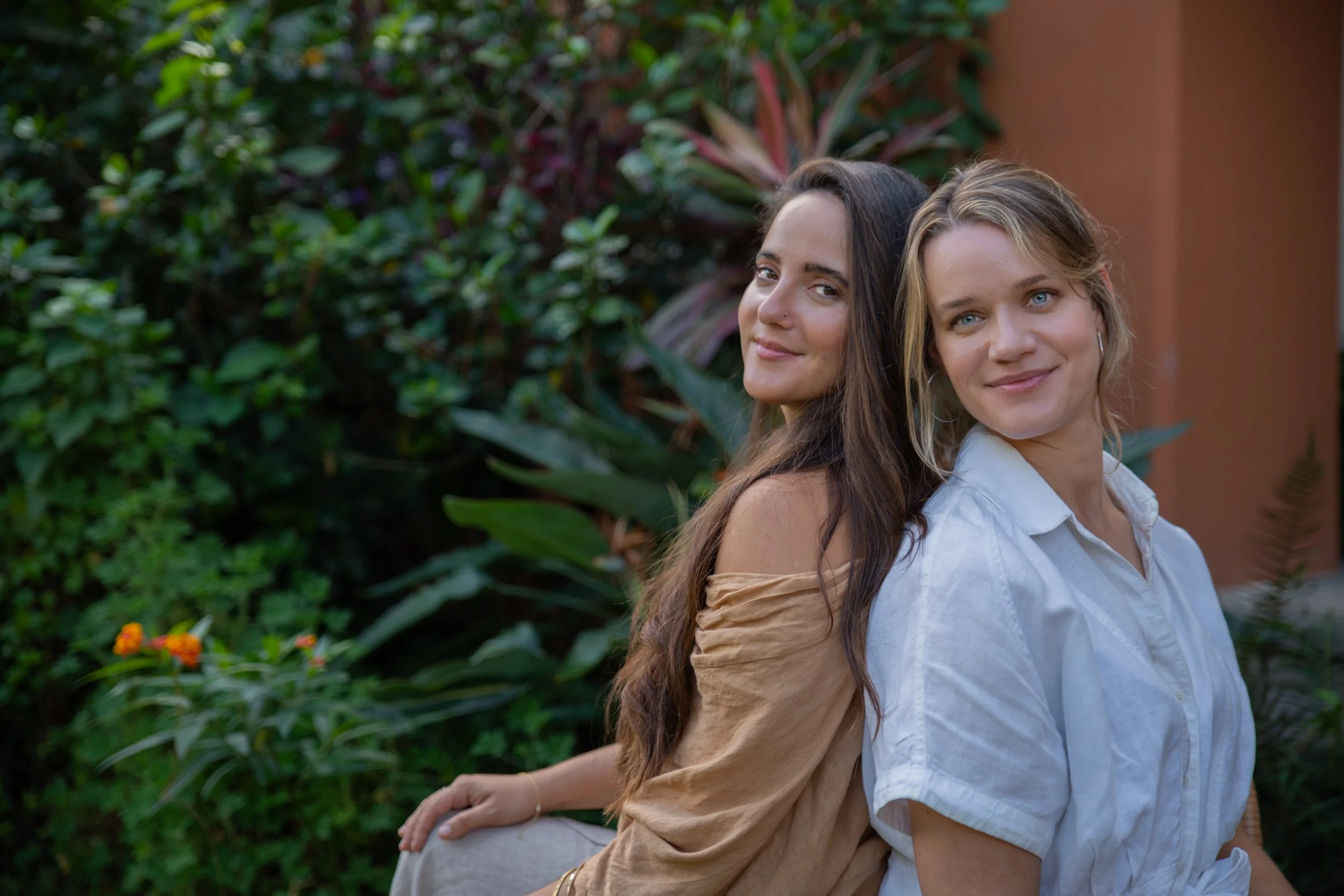 Two women sit back-to-back outdoors, smiling, surrounded by lush green foliage and plants.