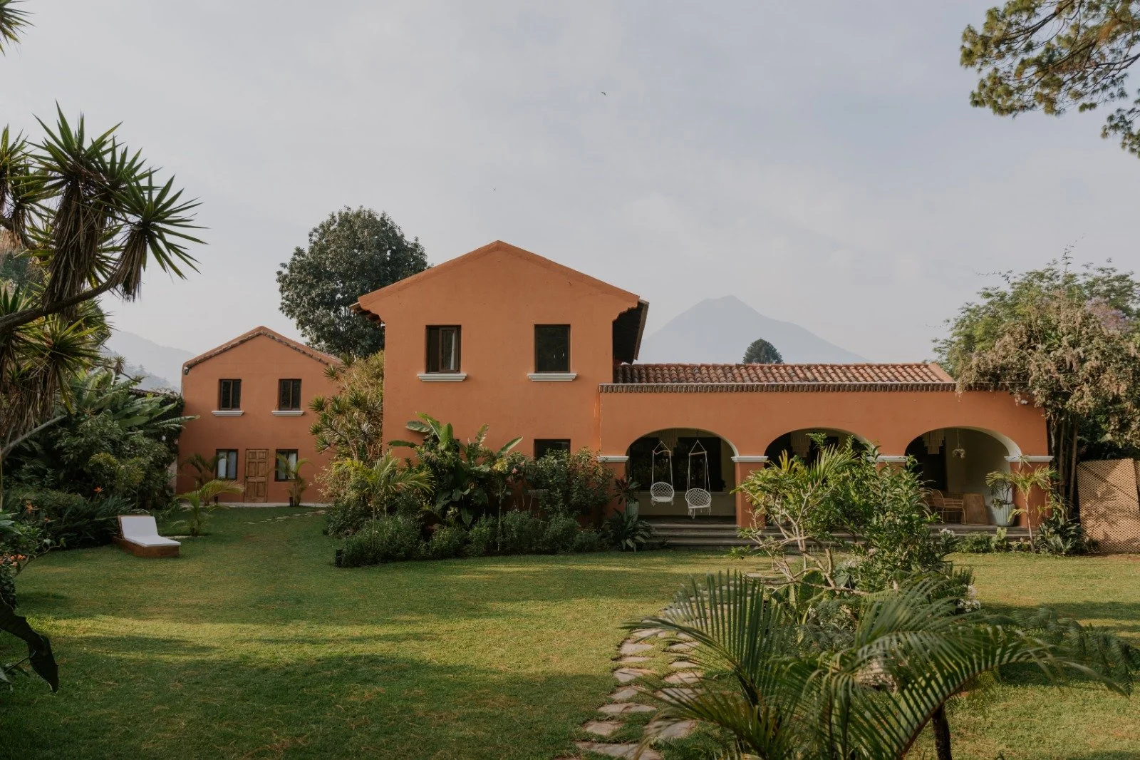 A house with peach-colored walls, a tiled roof, and arched patio doors, surrounded by lush green plants and trees, with lounge chairs and hanging chairs on the patio, under a cloudy sky with mountains in the background.
