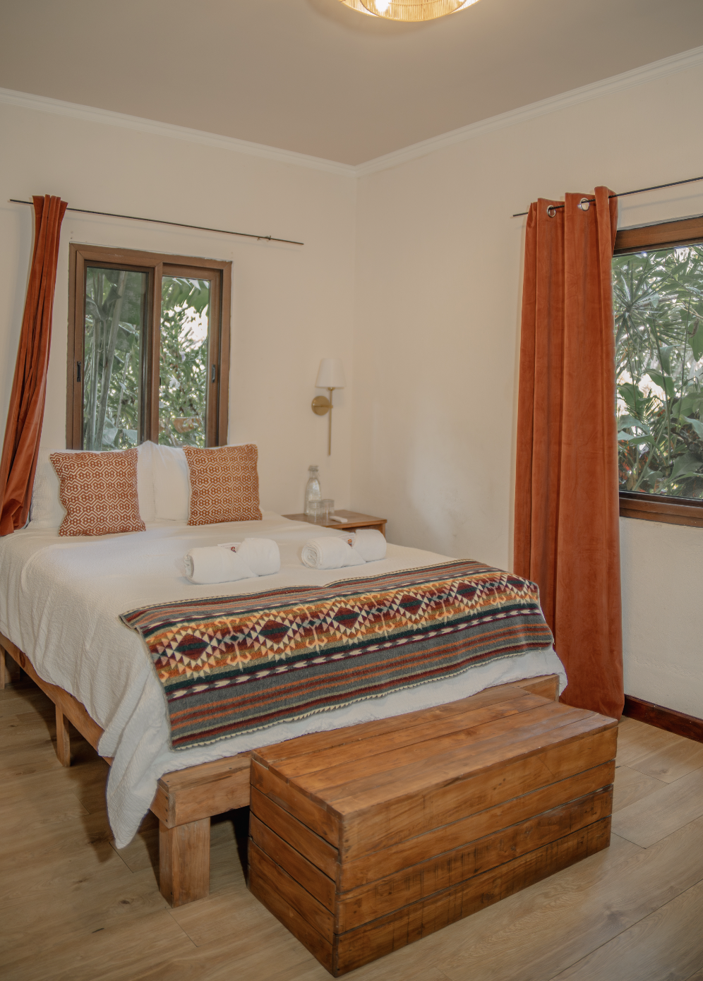 A bedroom with a bed decorated with two patterned pillows, a patterned blanket at the foot, and three rolled towels on top. There are two windows with orange curtains, a wooden chest at the foot of the bed, and a nightstand with a water bottle. The room has a wooden floor and white walls.