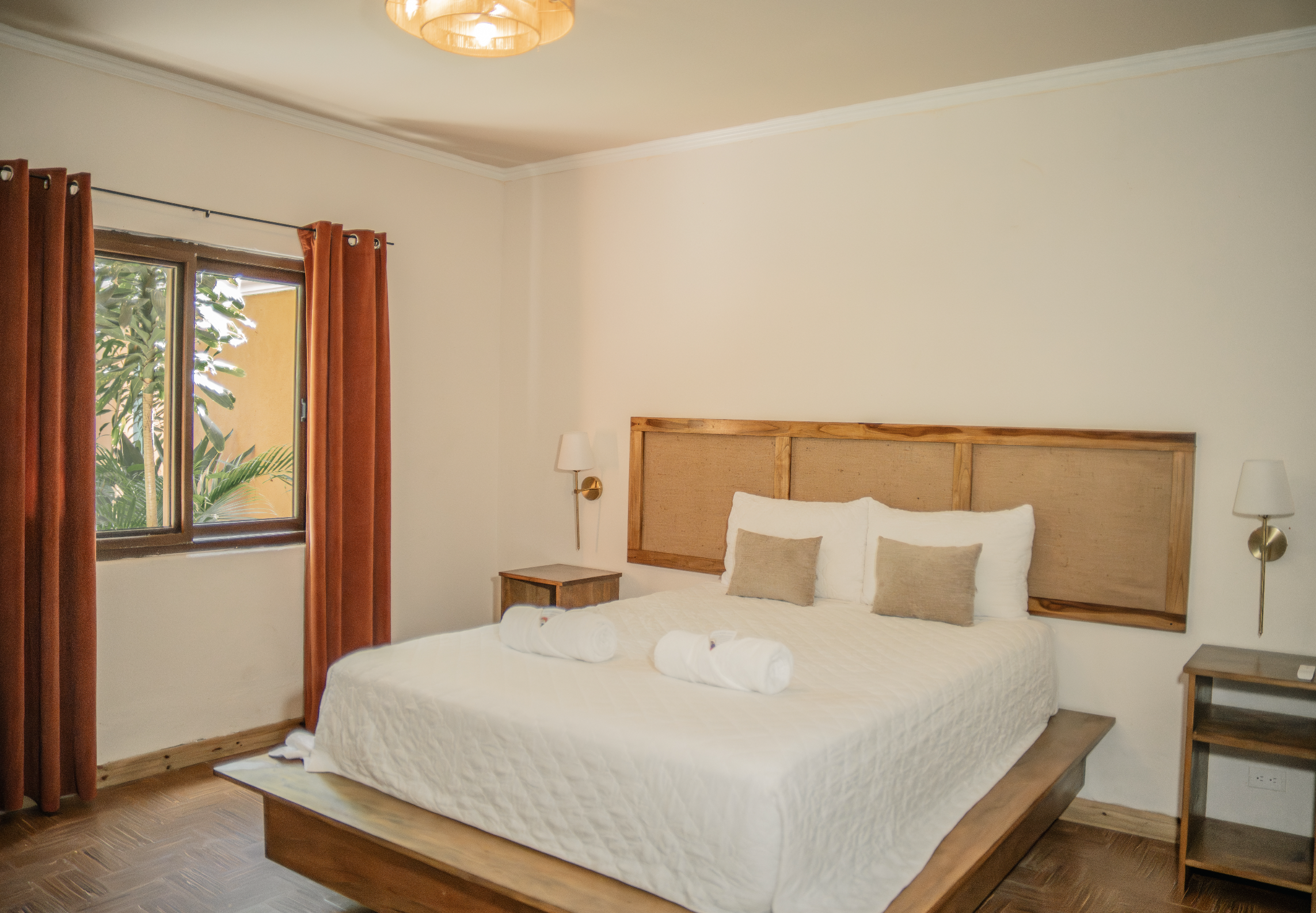 A bedroom with a large bed, wooden headboard, white bedding, and beige pillows. There are two nightstands with lamps on each side of the bed. There's a window with orange curtains, and a plant can be seen outside the window. The room has wood flooring and a ceiling light fixture.