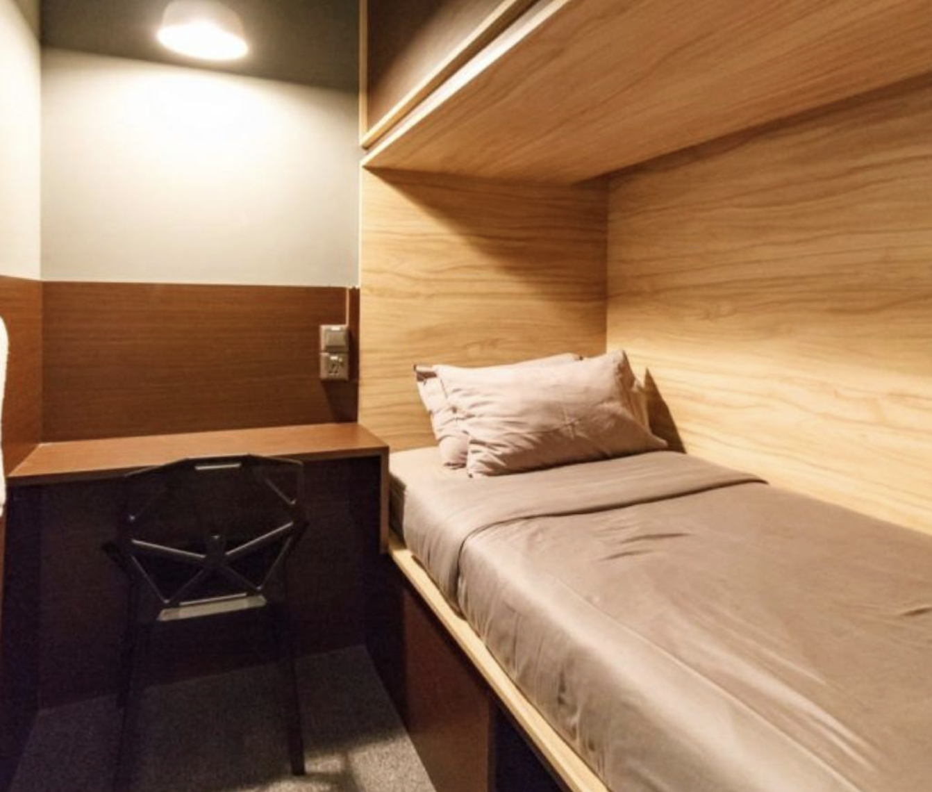 A cozy bedroom with a single bed, beige sheets, and a pillow, featuring wooden wall panels and a small desk with a black chair.