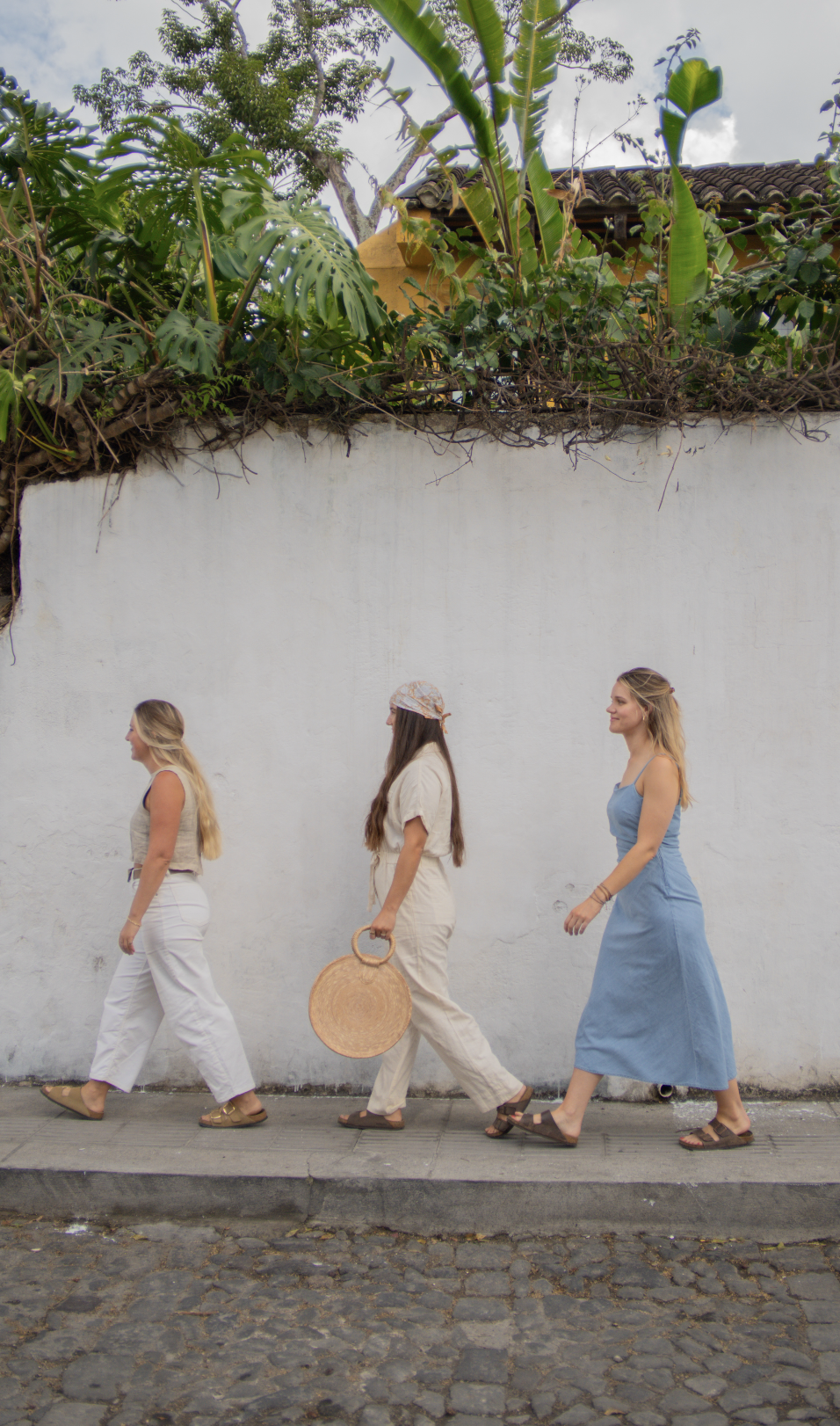 Three women walking along a sidewalk past a white wall with plants and trees above it in the background.