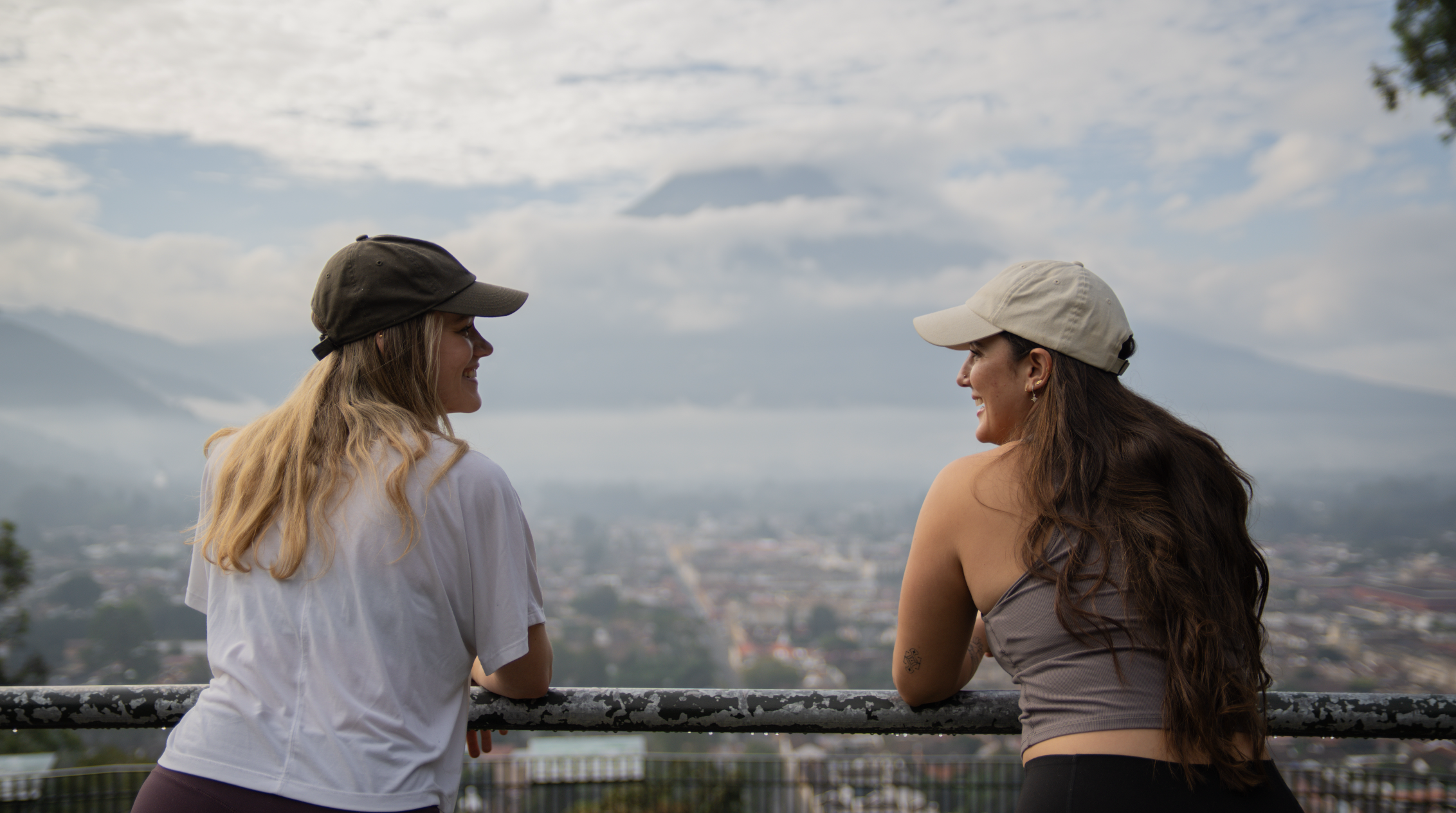 Two women wearing caps are smiling and talking over a railing, with a cityscape and mountains in the background.