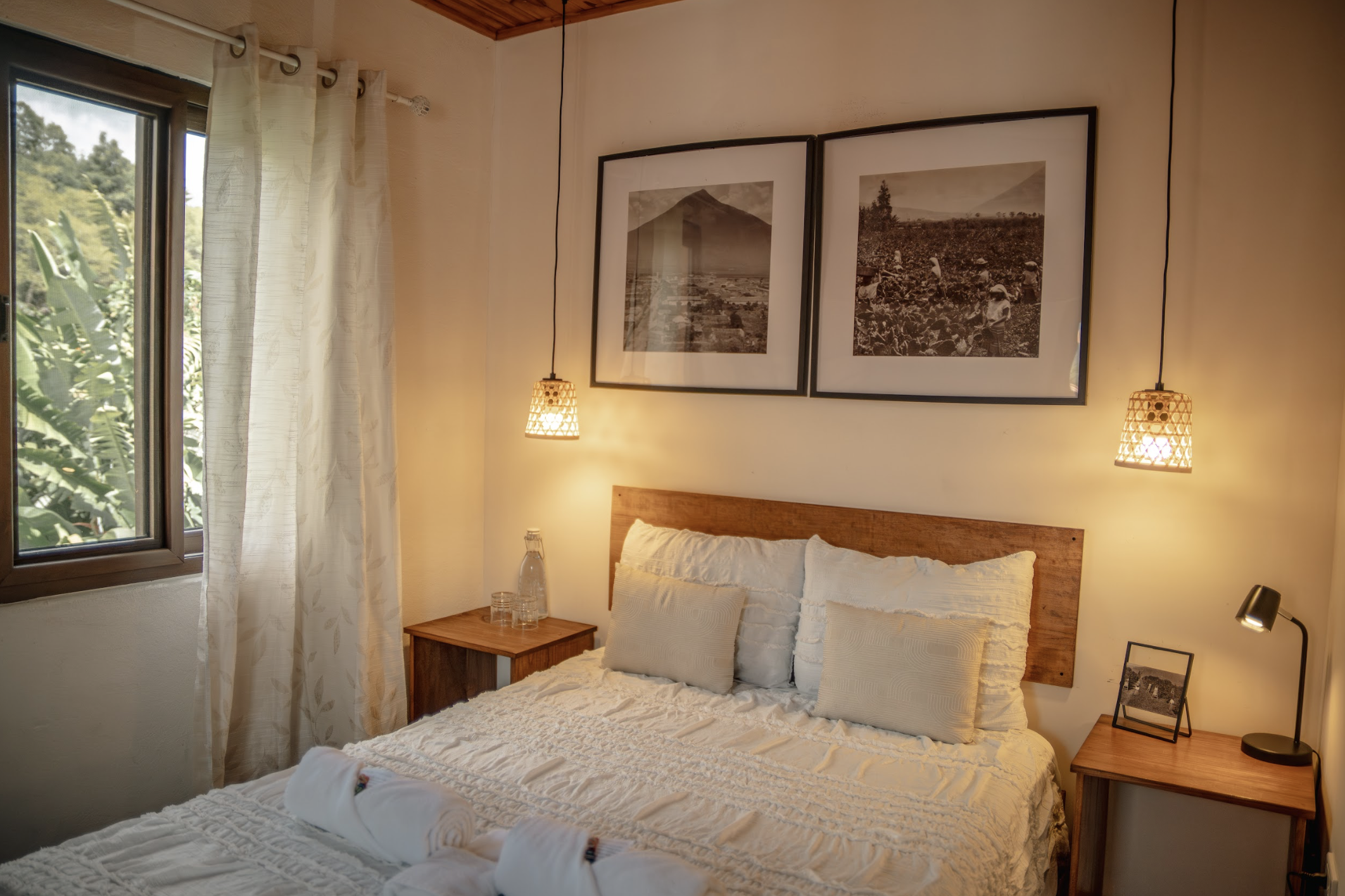Bedroom with two wooden nightstands, two beige pillows, and a bed with white bedding. Framed black and white photographs hang above the bed. Two pendant lights and a small desk lamp illuminate the room. A window with white curtains shows greenery outside.
