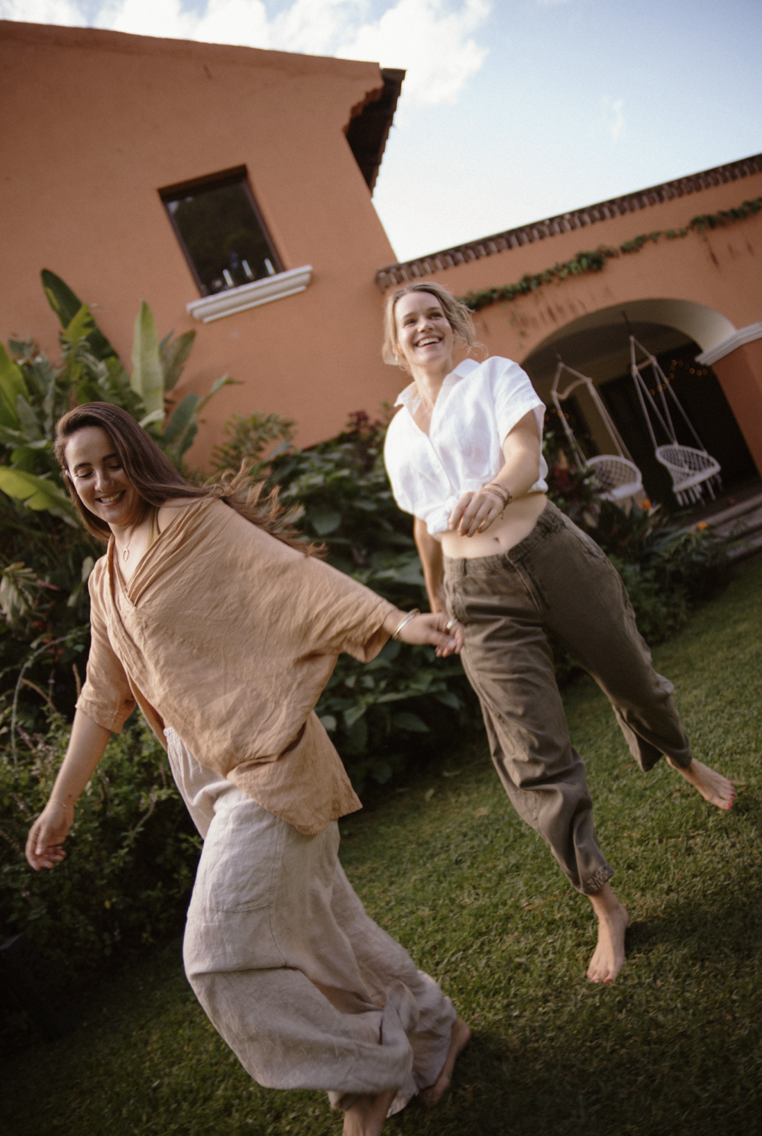 Two women are dancing and holding hands in a garden in front of a house with an orange exterior, greenery, and outdoor furniture, smiling and enjoying the moment.