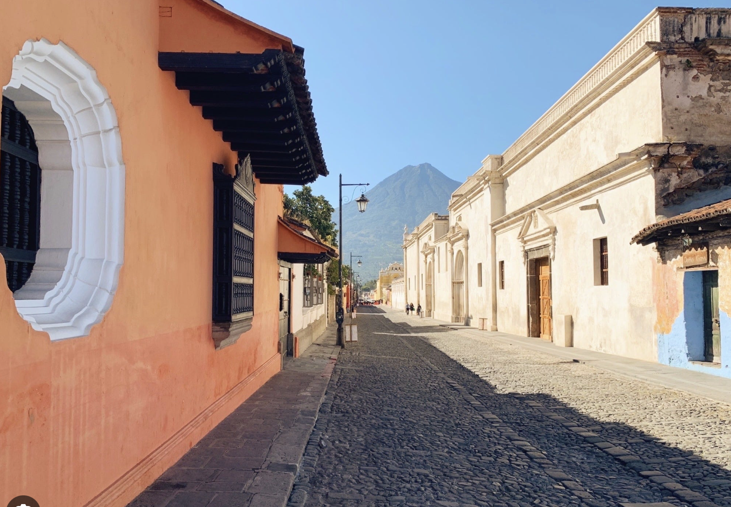 A cobblestone street lined with colorful old buildings in a historic town, with a prominent mountain in the background and a clear blue sky.