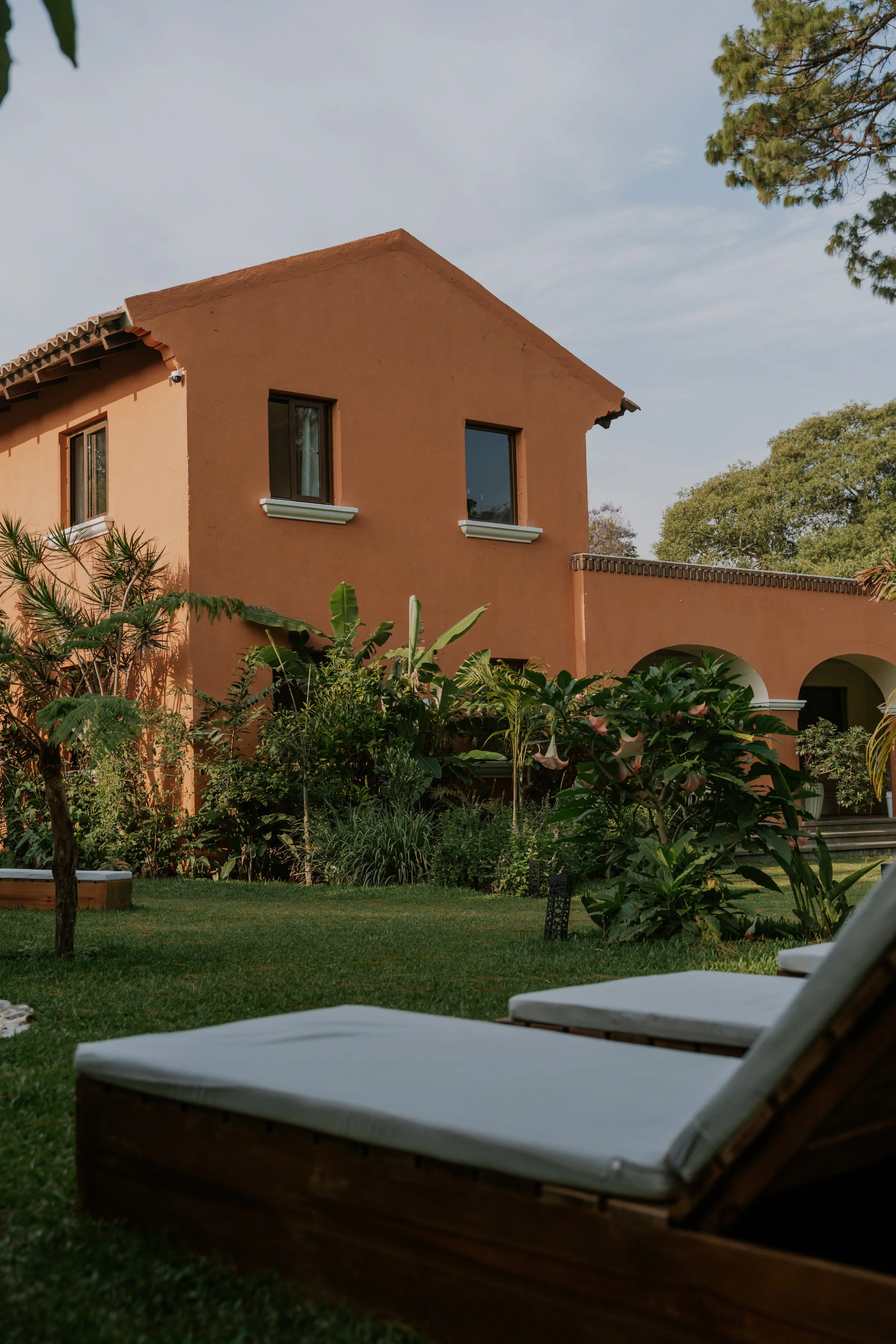 A peach-colored house with a garden in the foreground, lounge chairs on the lawn, and trees against a blue sky.