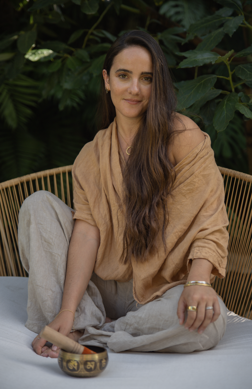 A woman with long brown hair sitting on a cushion, holding a Tibetan singing bowl with a wooden striker. She is wearing beige linen clothes and jewelry, and is surrounded by green foliage.