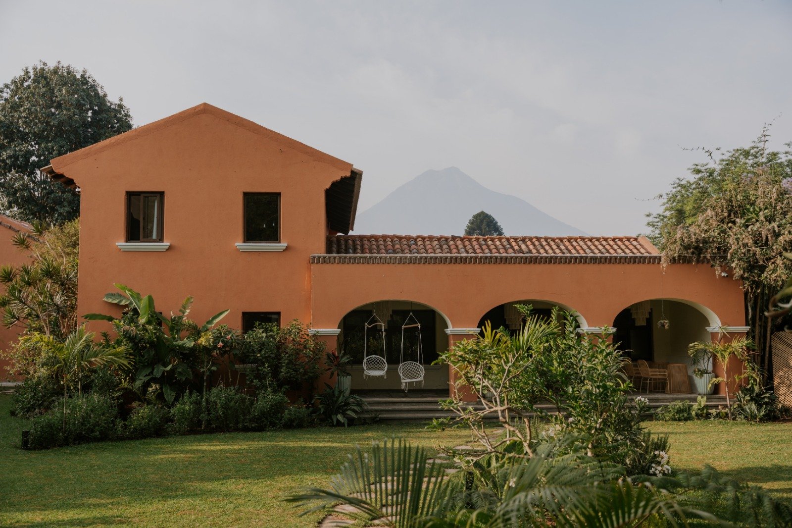 Orange two-story house with tiled roof, surrounded by lush green plants and trees, with a mountain visible in the background.