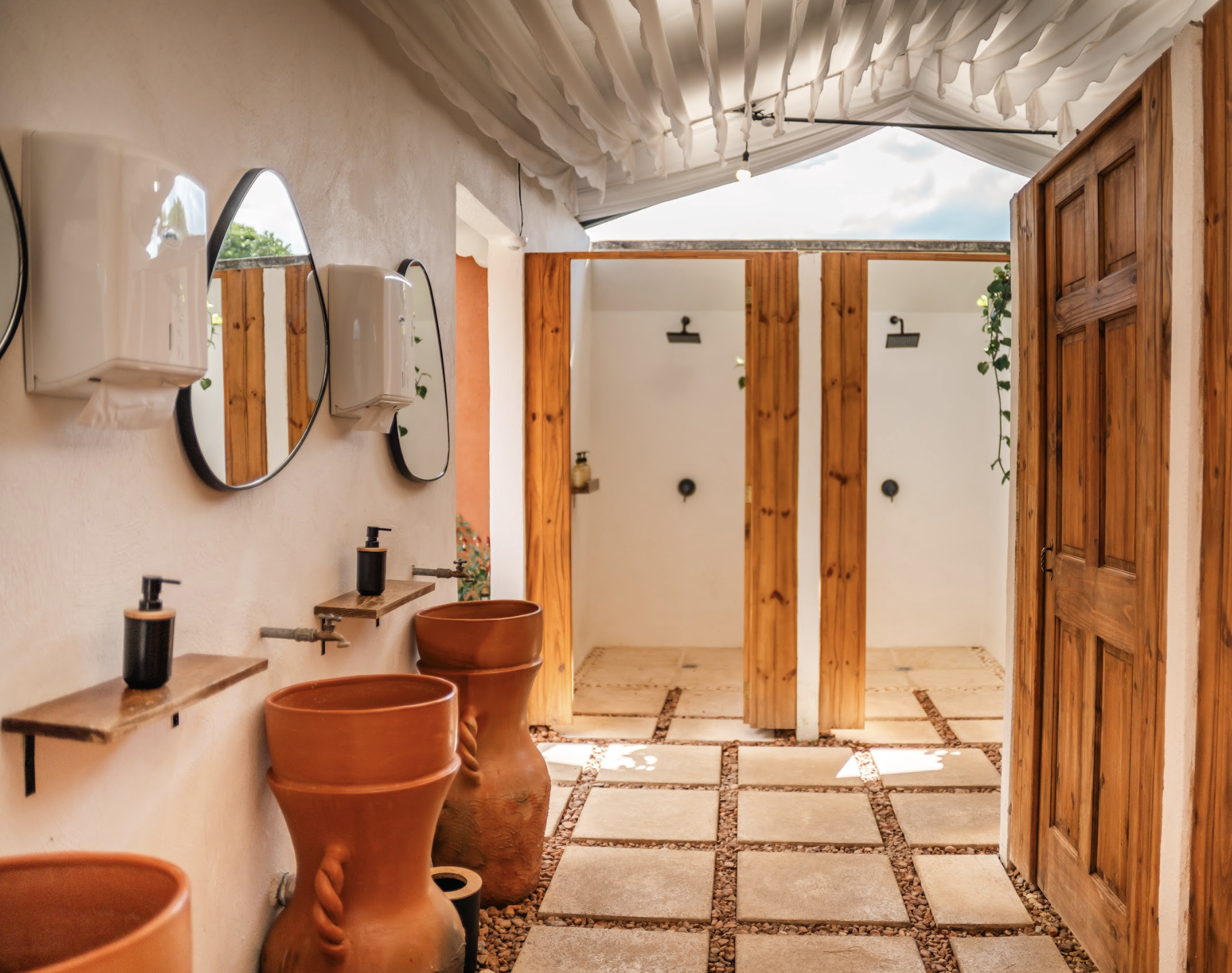 Public outdoor shower area with multiple shower stalls, wooden doors, and terracotta sinks with mocha liquid soap dispensers, set in a lush tropical setting.