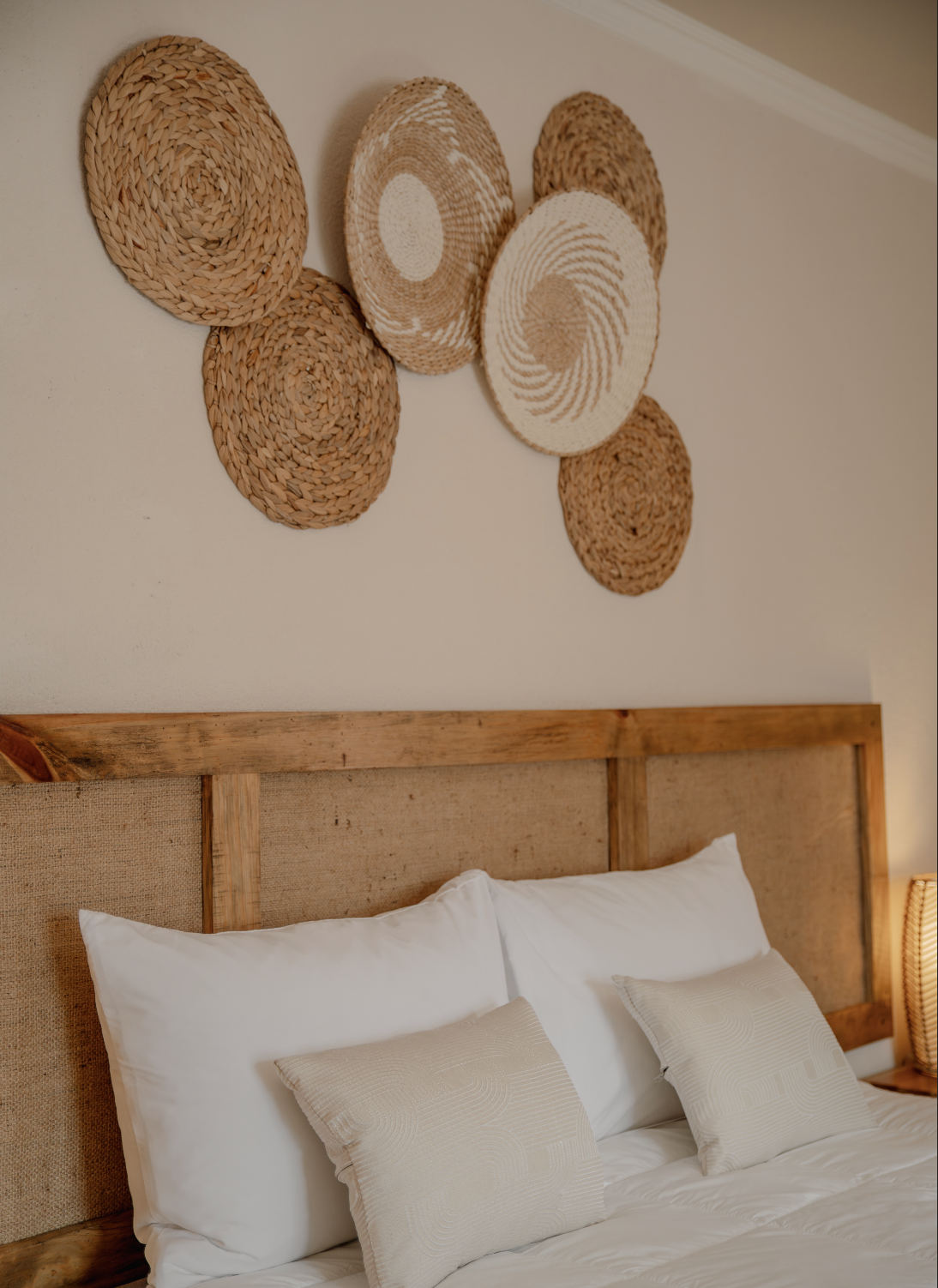 Decorative woven wall art above a bed with white pillows and beige throw pillows in a bedroom with a wood headboard and bedside lamp.