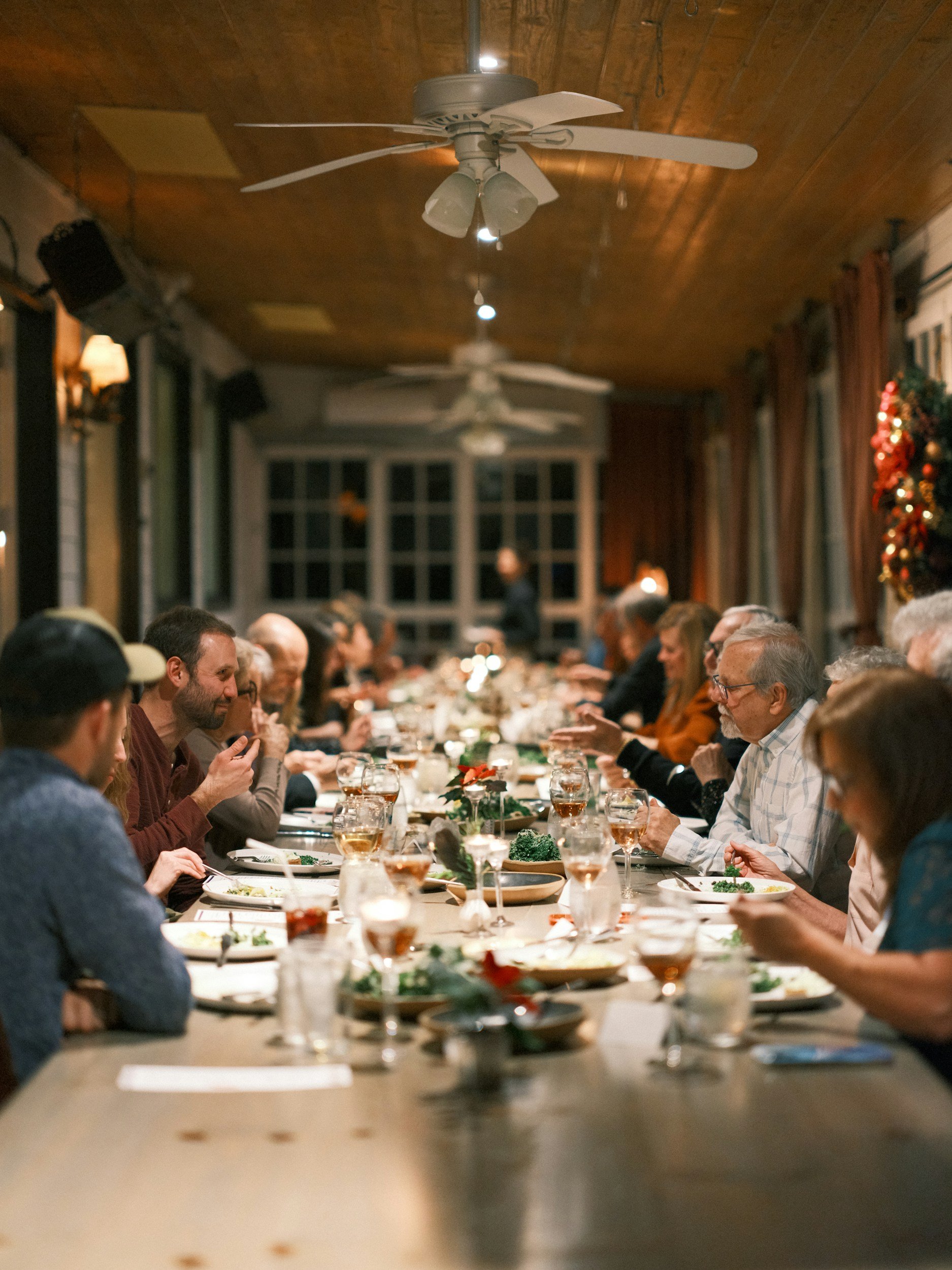 People dining at a long, decorated holiday dinner table in a cozy room with wooden ceiling and Christmas decorations.