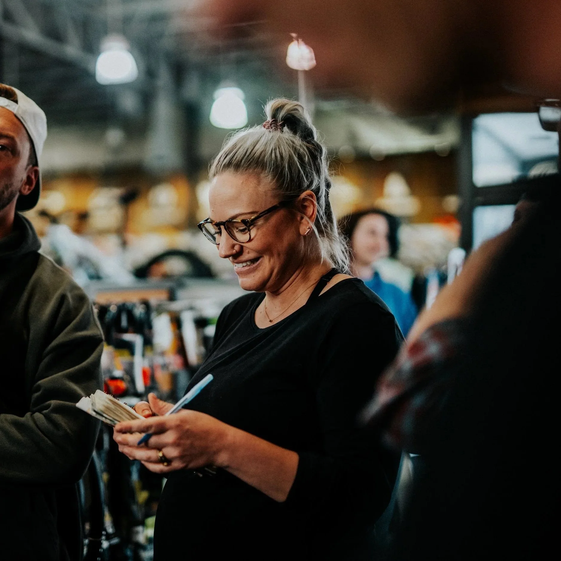 A woman with glasses and blonde hair in a ponytail smiling while writing in a notebook in a busy indoor setting.