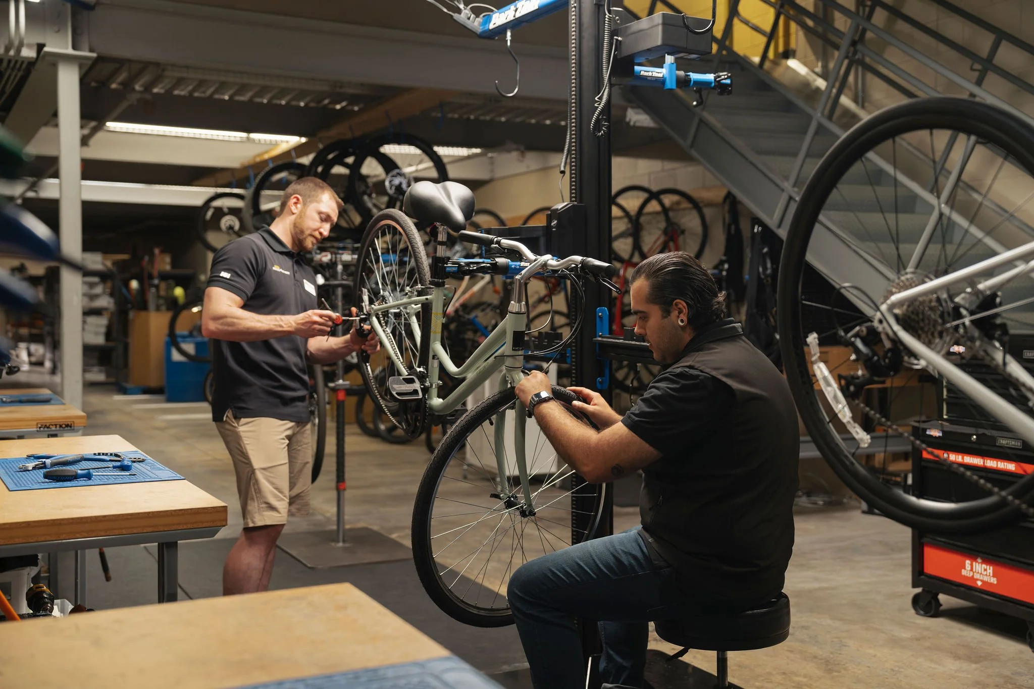Two men assembling or repairing a mountain bike inside a bike shop, with multiple bikes on racks and workbenches around them.