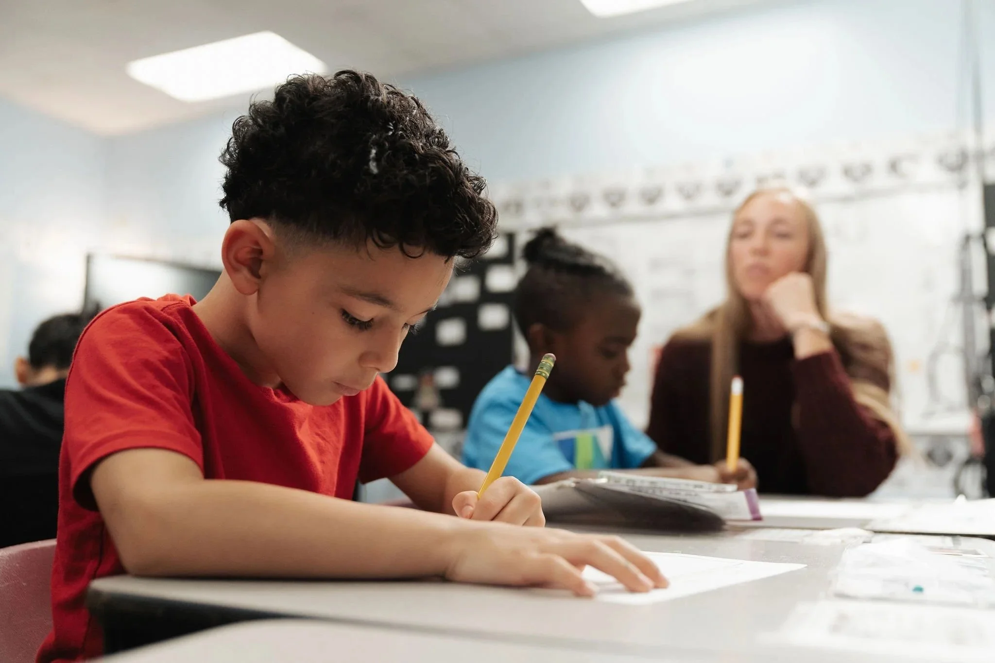Children sitting at desk, taking notes, with a teacher observing in the background.
