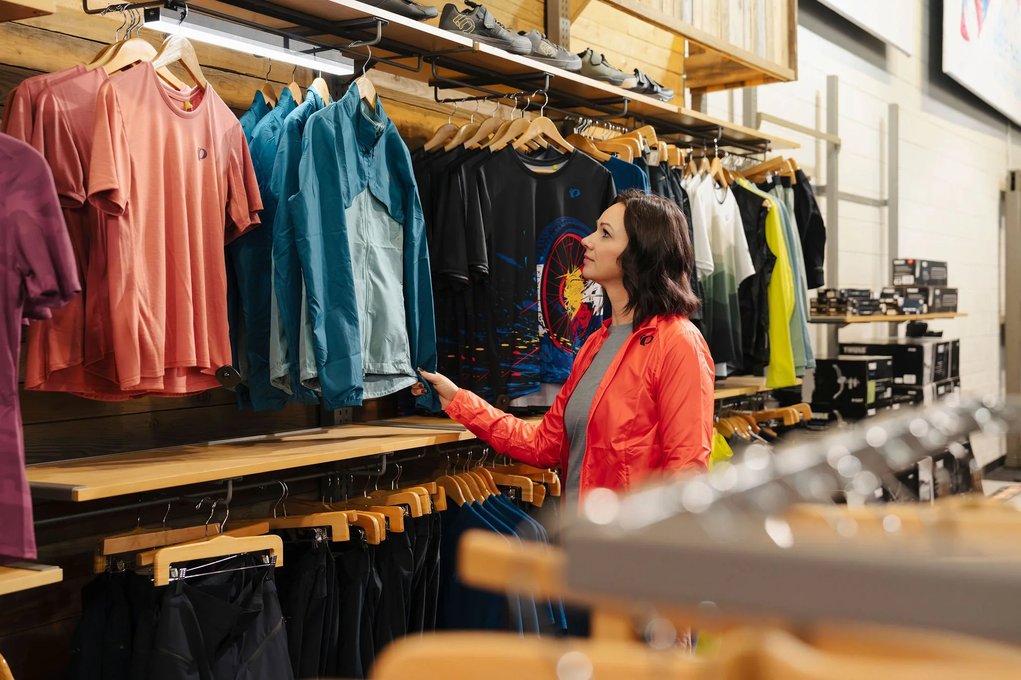 A woman shopping for athletic clothing, browsing a display of colorful sports t-shirts, jackets, and pants in a retail store with wooden shelves and hanging racks.