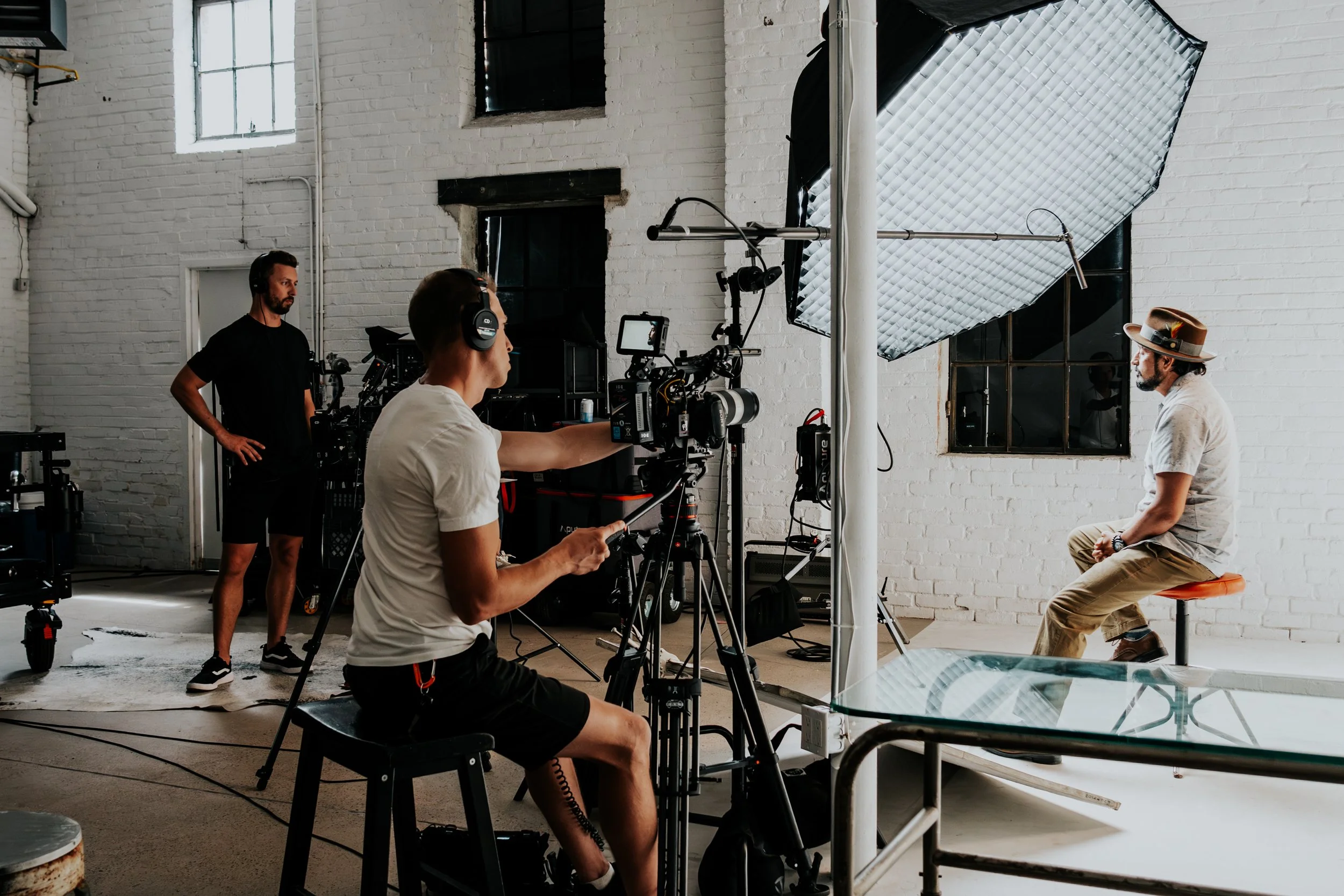 A man wearing a hat, sitting on a stool, being filmed in a studio with white brick walls, large window, and professional lighting equipment.