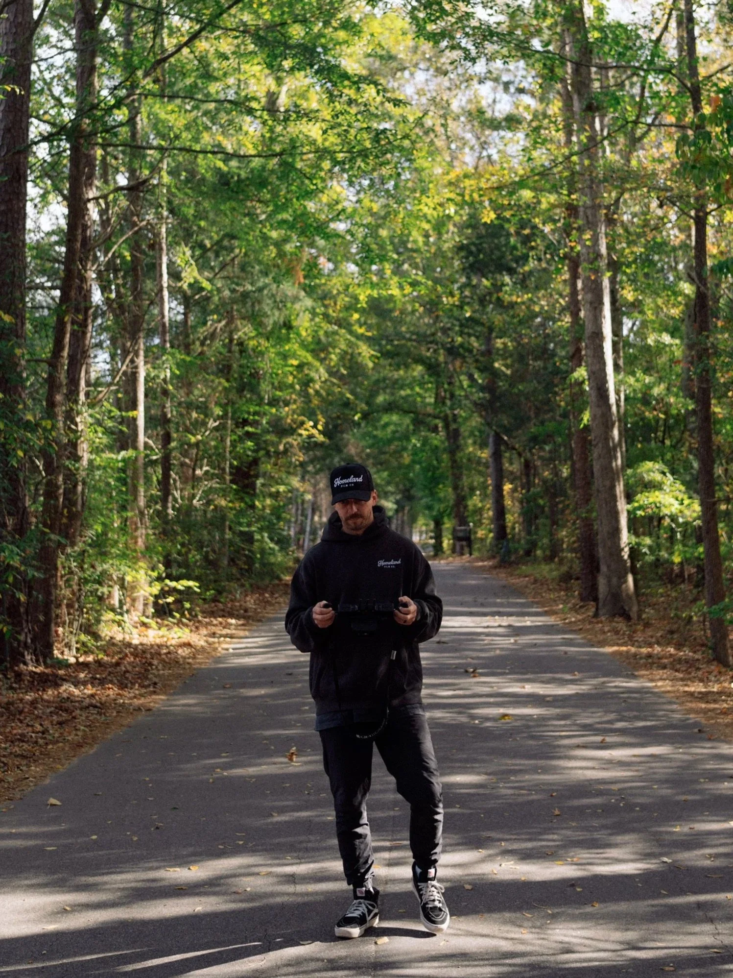 A man wearing a black hoodie, black pants, and sneakers, holding a drone remote control, standing on a paved path in a sunlit forest with green trees.