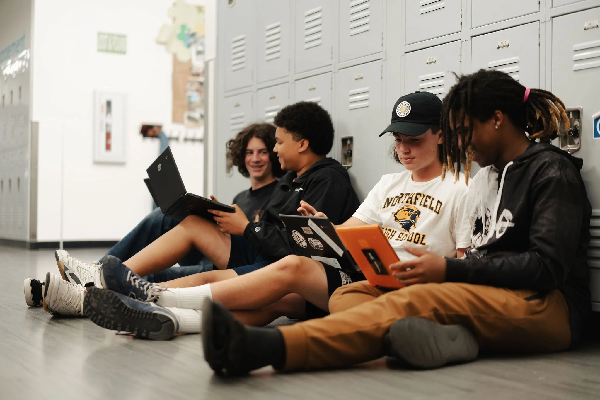 Four students sitting on the floor against lockers, using electronic devices and chatting.