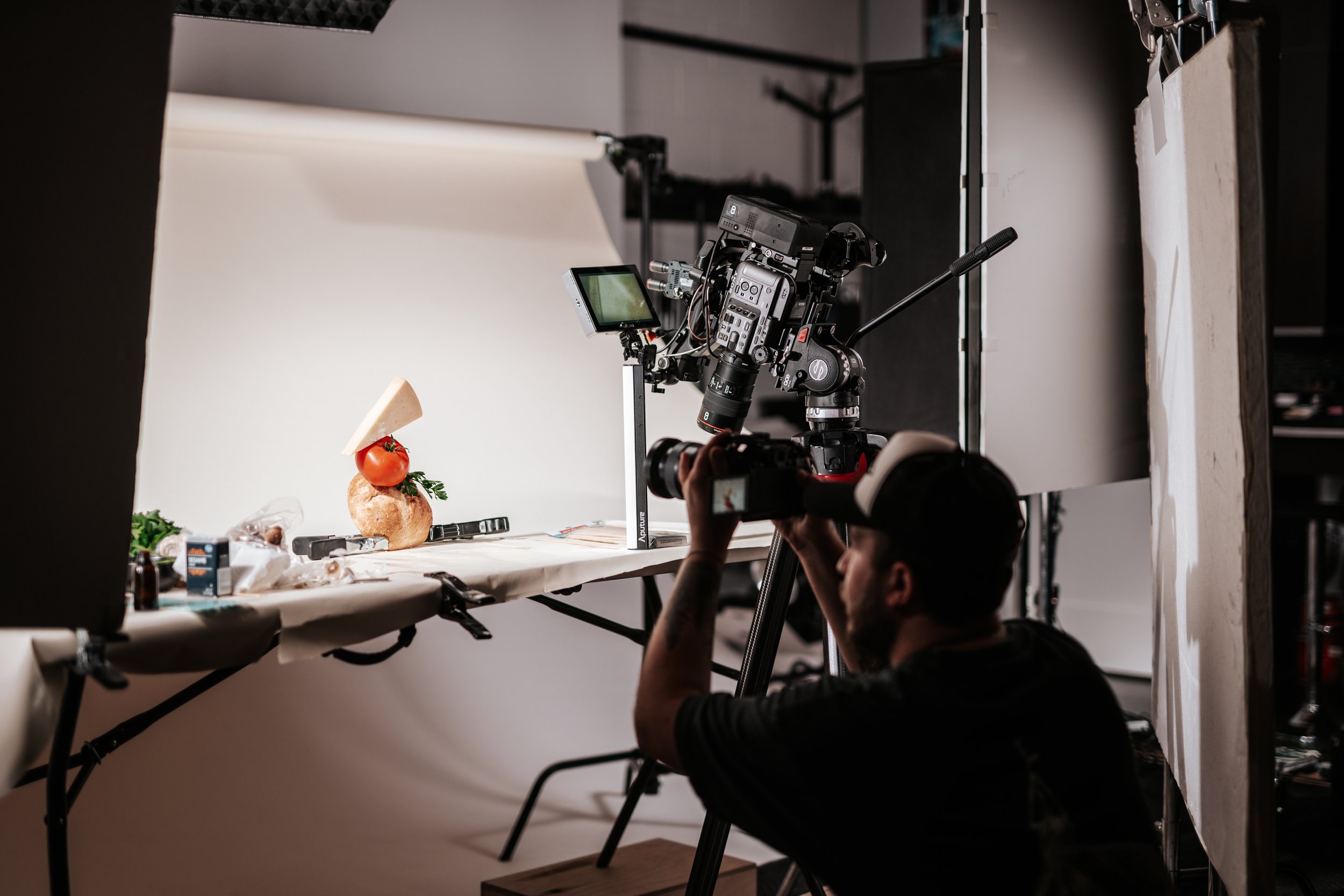 A photographer is filming a food tableau featuring a tomato, a mushroom, a piece of cheese, and some greens in a studio setting with professional lighting and backdrop.