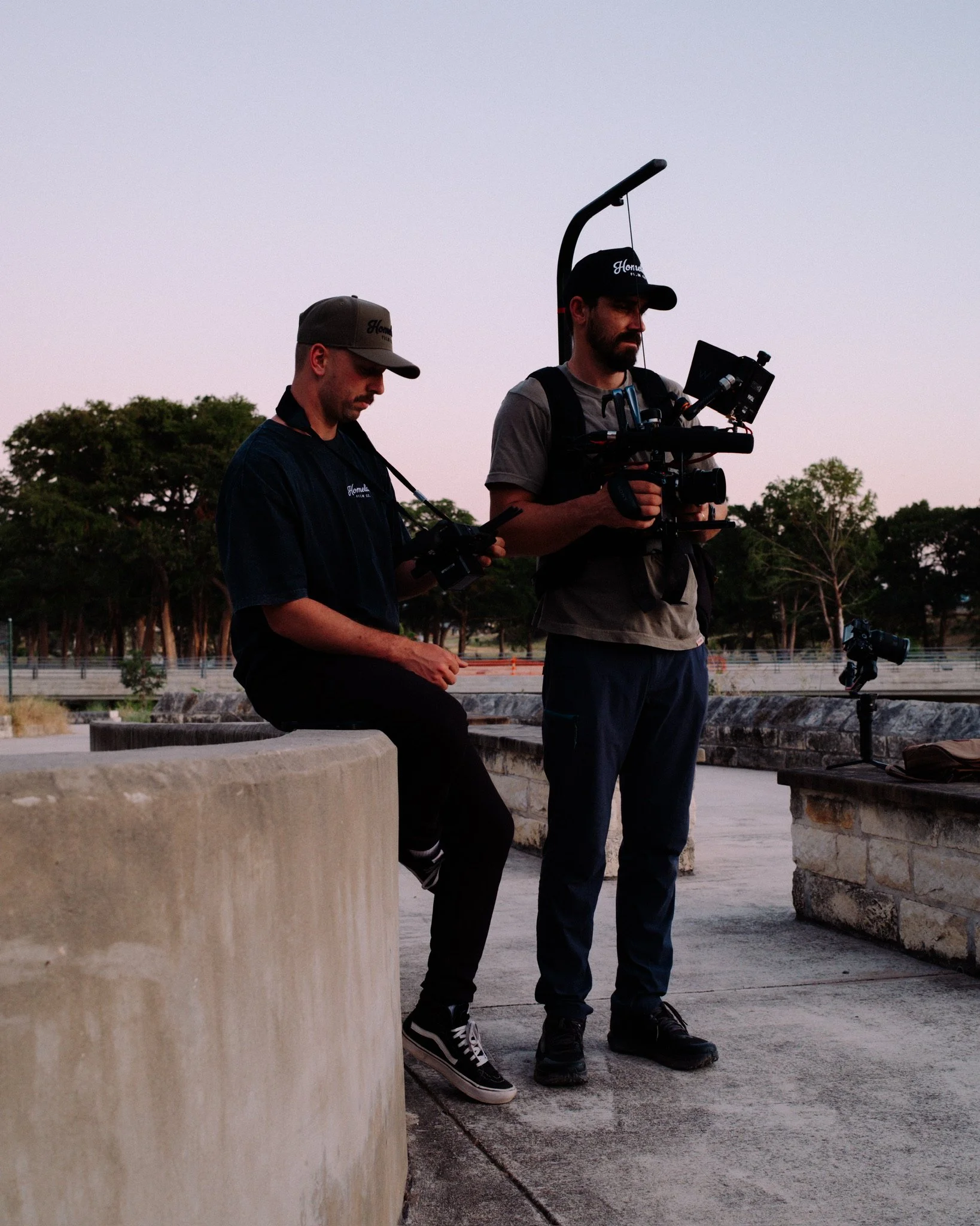 Two men operating camera equipment outdoors near a concrete barrier with water and trees in the background during sunset or dusk.
