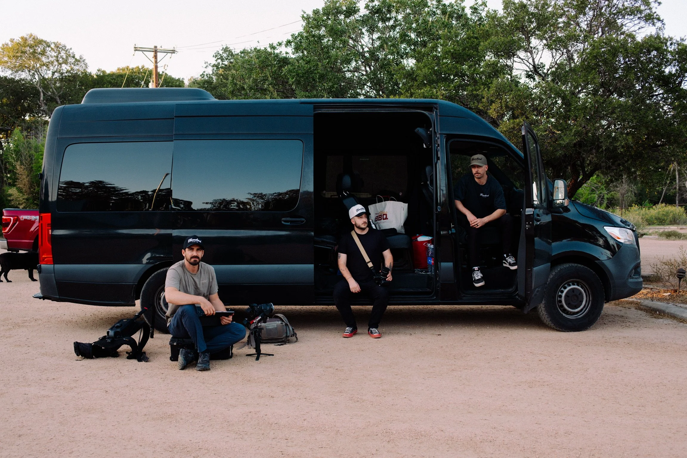 Three men sitting and standing near a black van outdoors with trees in the background. One man is sitting on a small black case, another is sitting on the van's open sliding door with a camera hanging from a strap around his neck, and the third man is sitting on the van's front seat with the door open. Equipment and bags are on the ground beside them.
