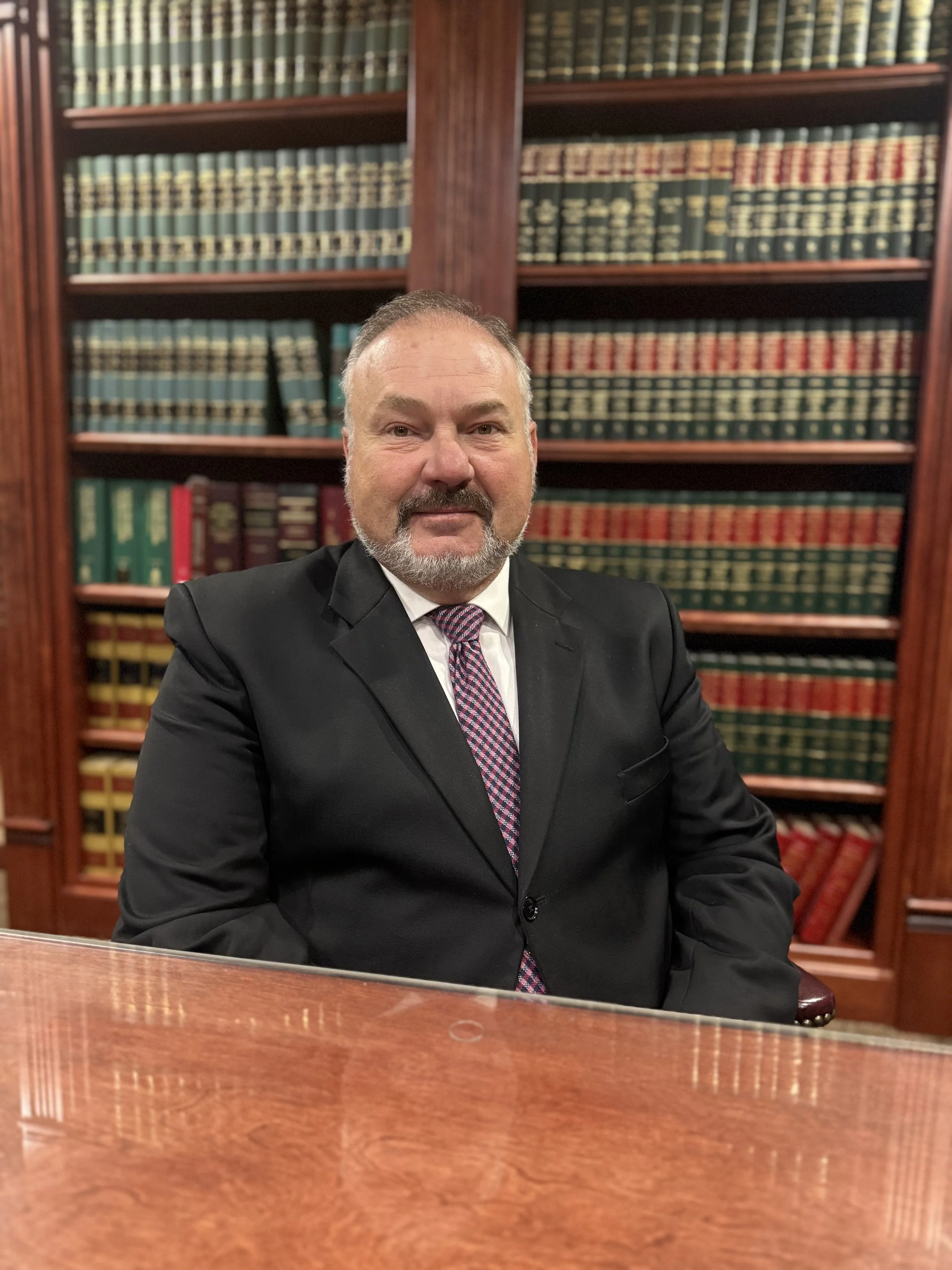 A man with a beard and mustache wearing a black suit, white shirt, and multicolored tie, sitting at a wooden table in a library with shelves of books in the background.