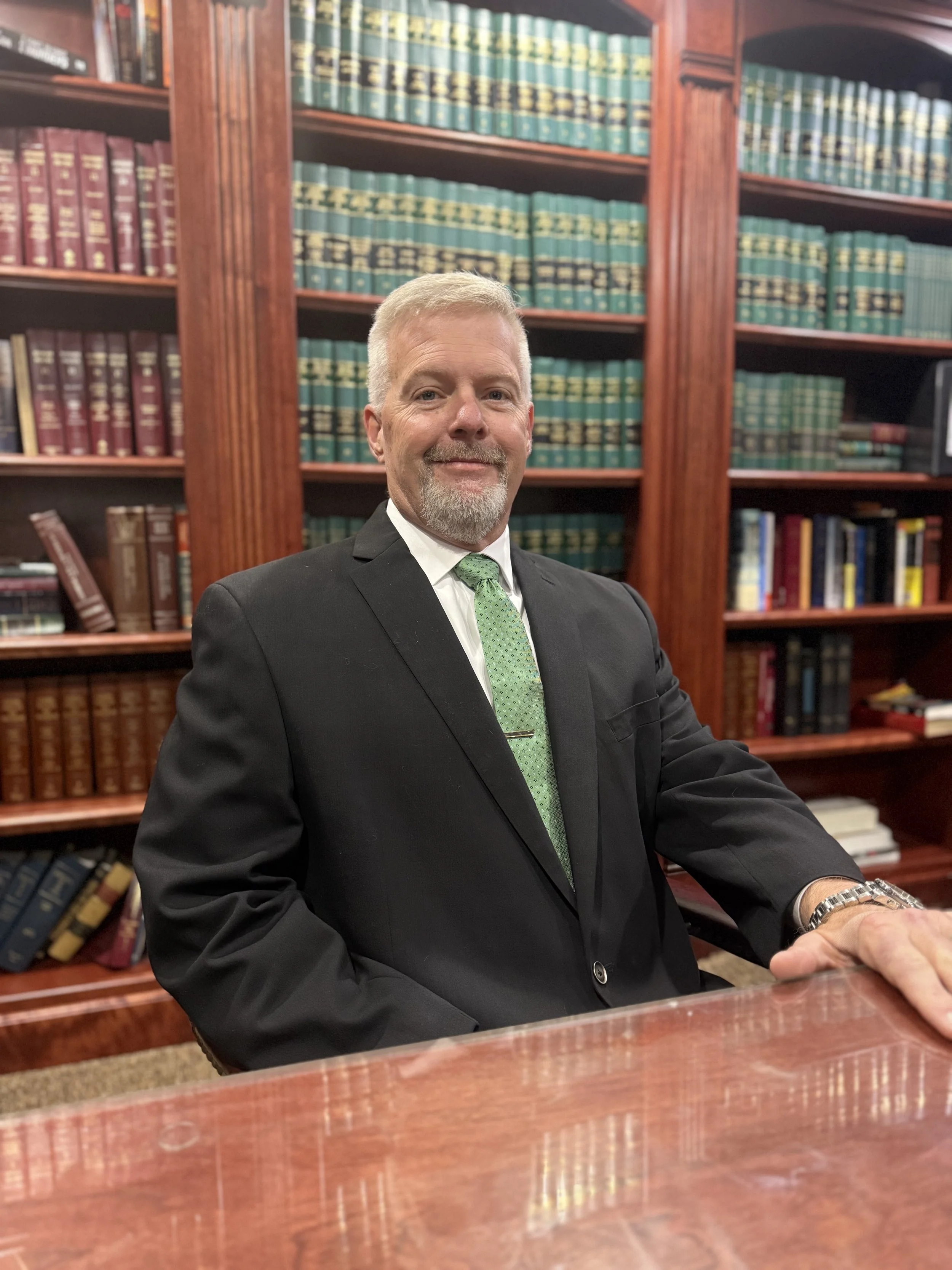 A middle-aged man with grey hair and a beard wearing a black suit, white shirt, and green tie, sitting at a wooden desk in a library filled with leather-bound books.