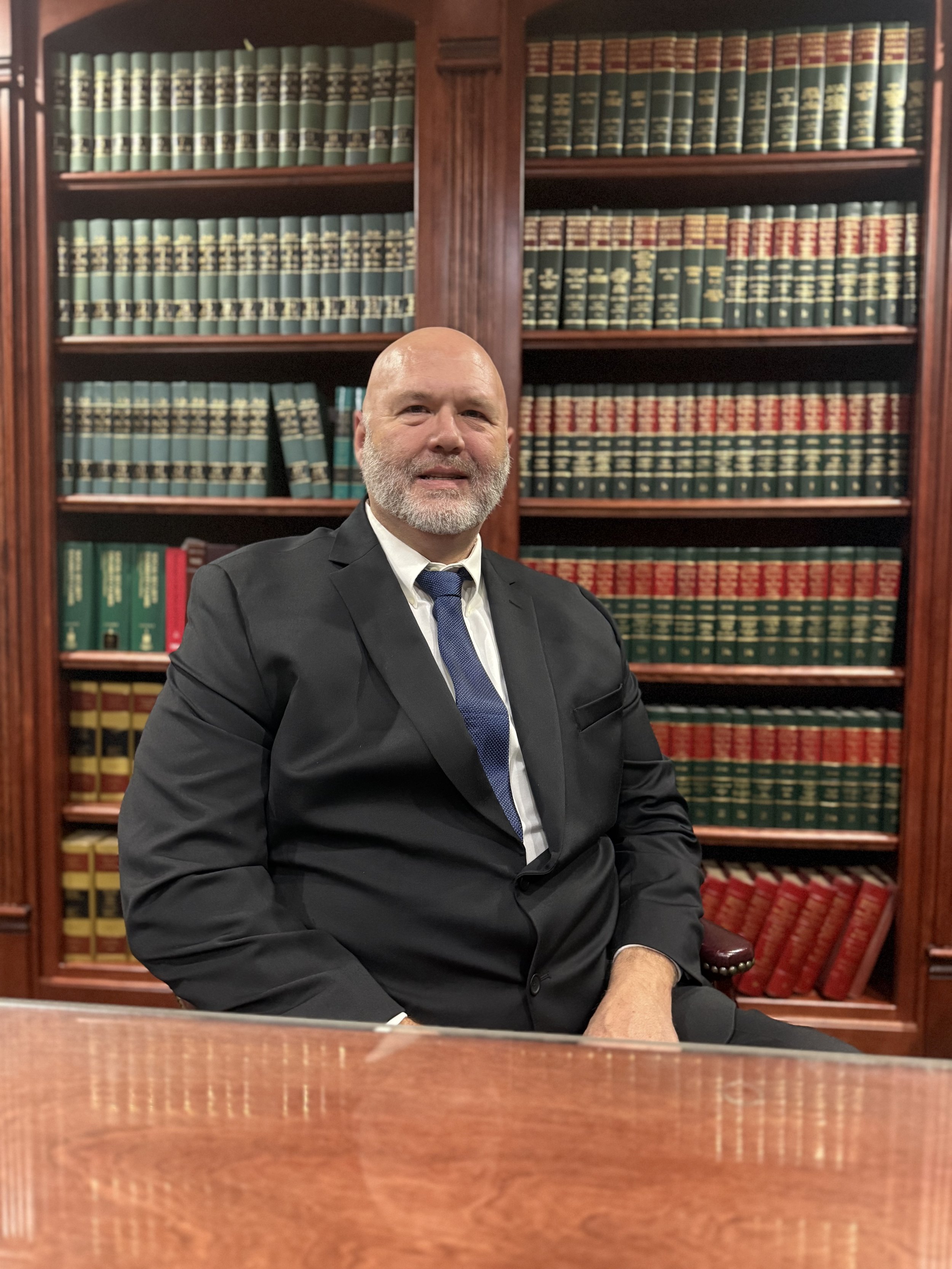 A man with a beard in a suit and tie, sitting in a lawyer's office with wooden bookshelves filled with law books in the background.