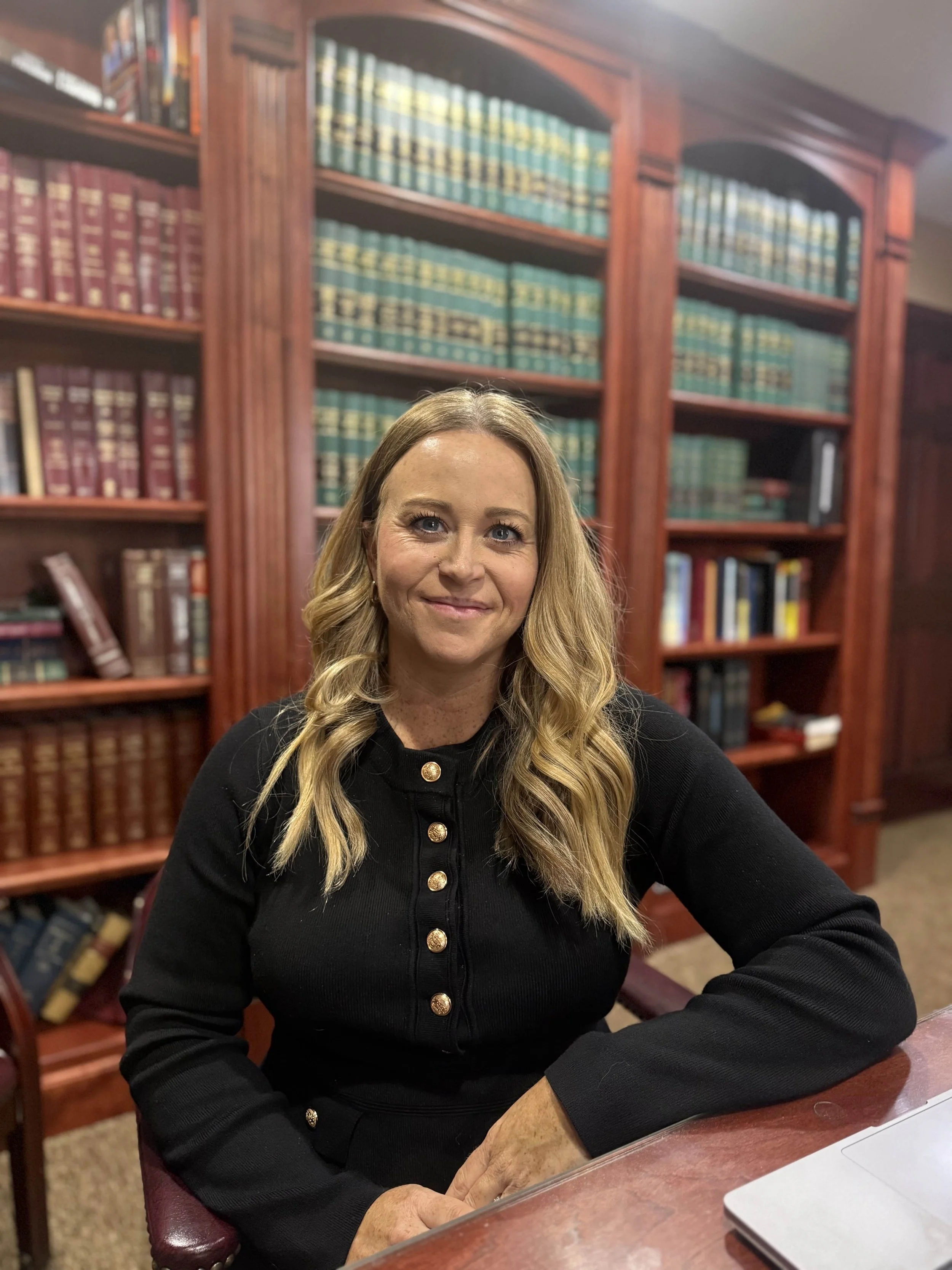 A woman with long blonde hair, wearing a black top with gold buttons, sitting at a wooden desk in a library with large wooden bookshelves filled with legal or reference books in the background.