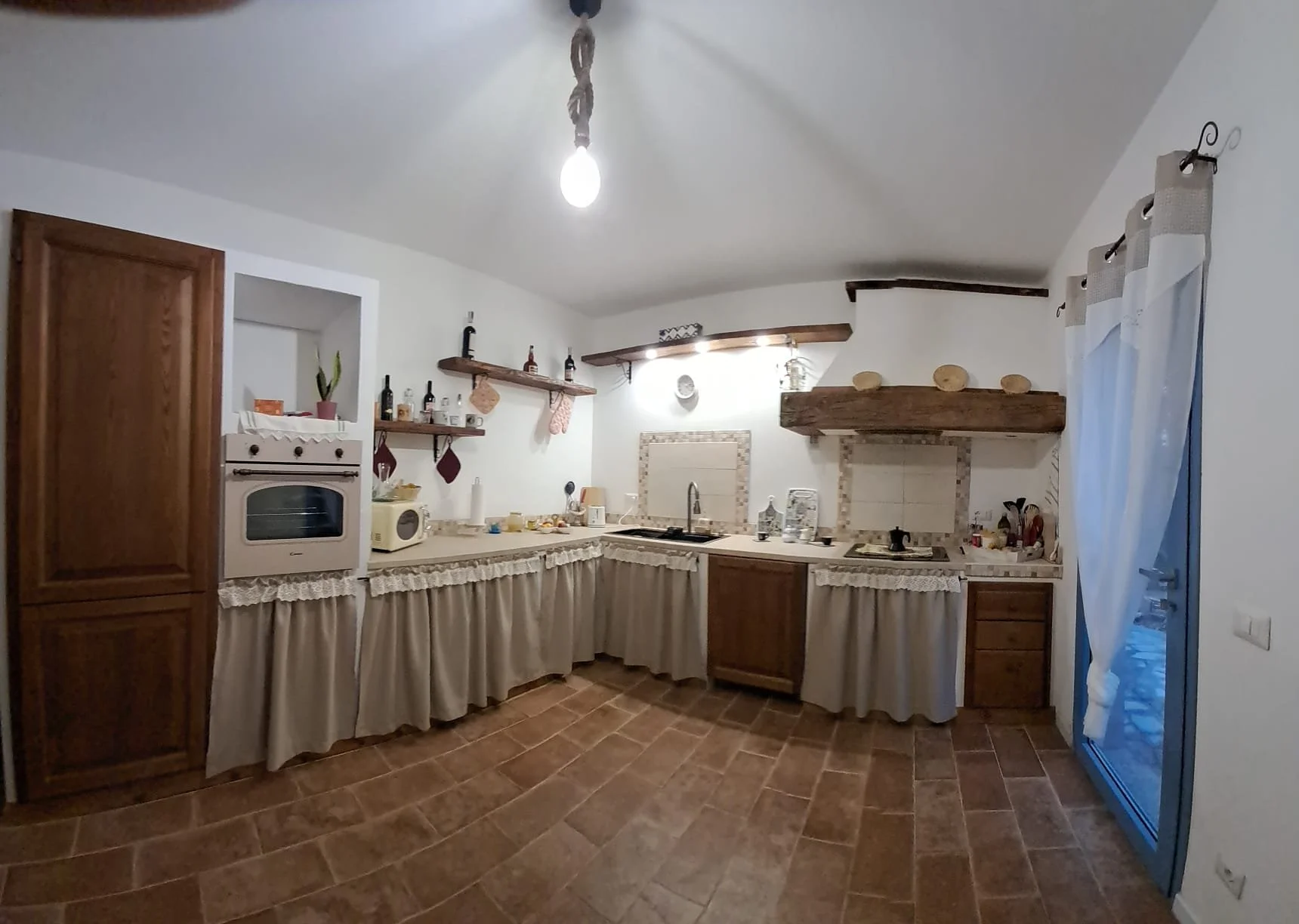 Cucina rustica con mobili in legno, piani di lavoro, scaffali con bottiglie e decorazioni, porta con tende bianche, illuminazione soffusa.