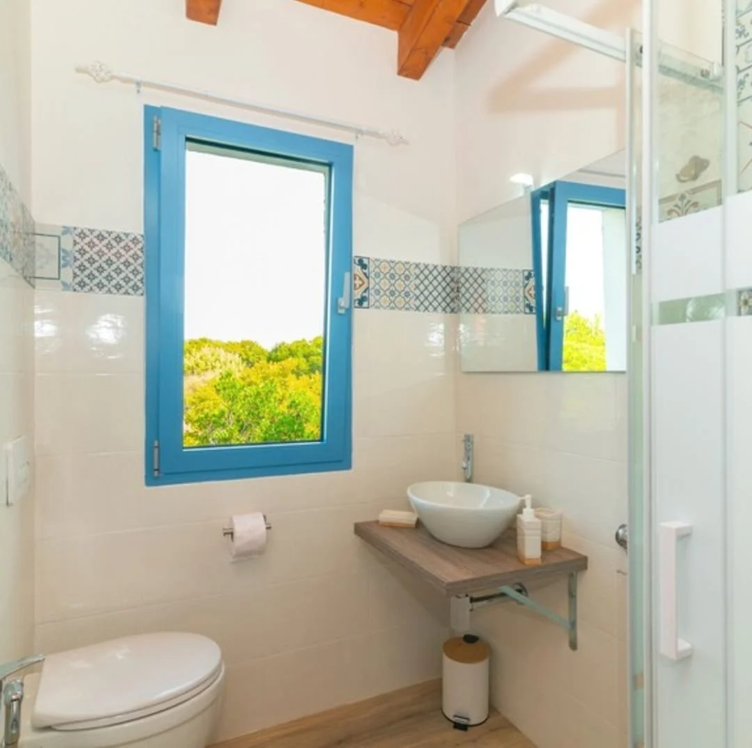 Bagno con finestra con vista su alberi verdi, lavandino sopra un piano in legno, specchio e servizi igienici