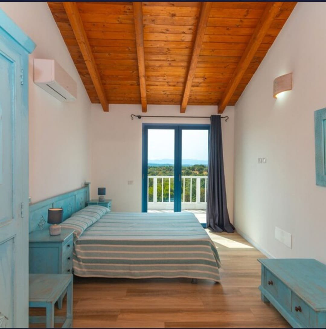 Camera da letto con letto matrimoniale con coperta a righe blu e bianca, mobili in legno azzurro, pareti bianche, pavimento in legno, grande finestra con vista su paesaggio verde e balcone, lampade da muro, aria condizionata.
