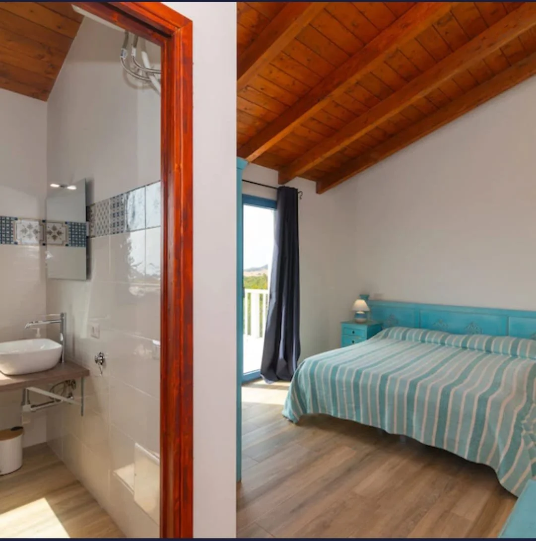 Camera da letto con soffitto in legno, letto con copriletto a righe azzurro e bianco, piccola lampada sul comodino, porta balcone aperta con vista su campagna, bagno annesso con lavabo e pasta decorativa sulle pareti.