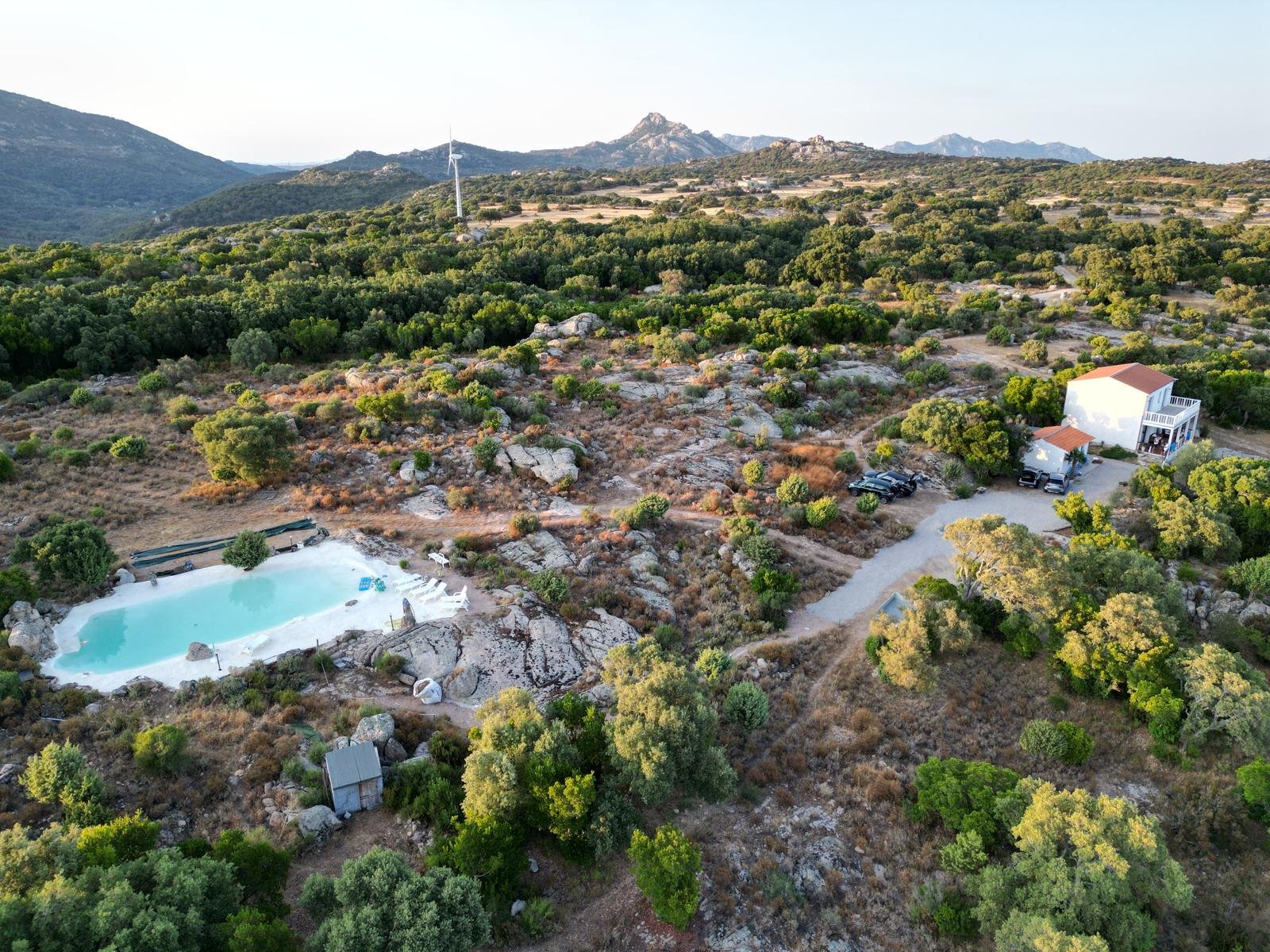 Paesaggio rurale con piscina, casa bianca con due piani, autovetture e vegetazione sulle colline con montagne sullo sfondo.
