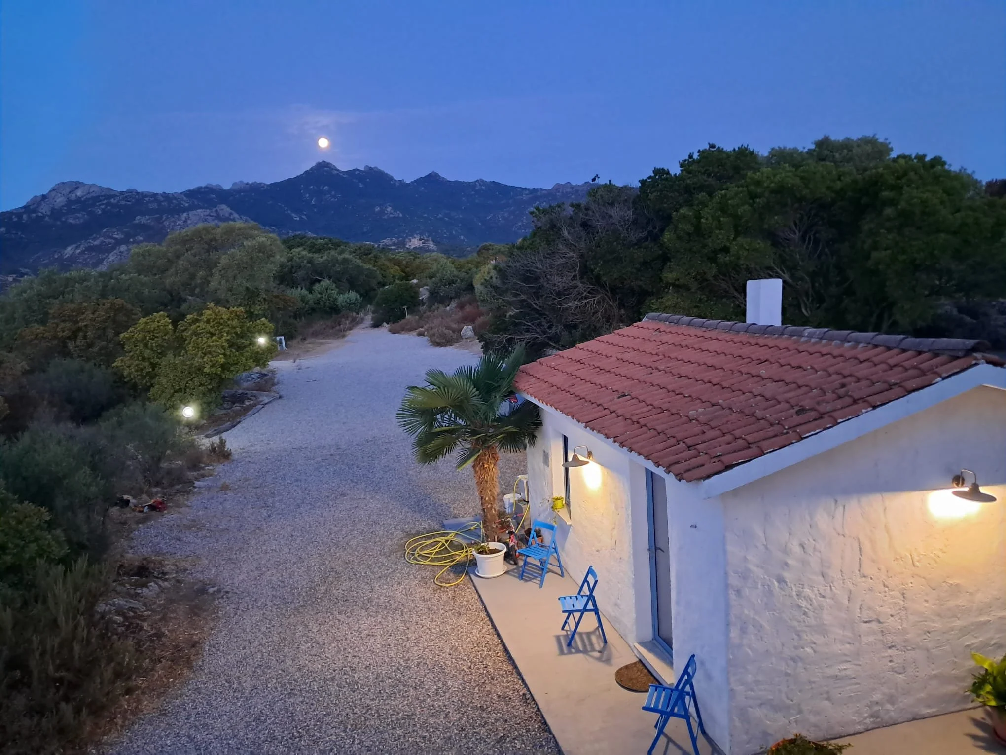 Casa bianca con tetto di tegole rosse, illuminata da luci esterne, circondata da piante e alberi, con un sentiero che si snoda verso le montagne sullo sfondo, sotto il cielo blu con la luna piena.