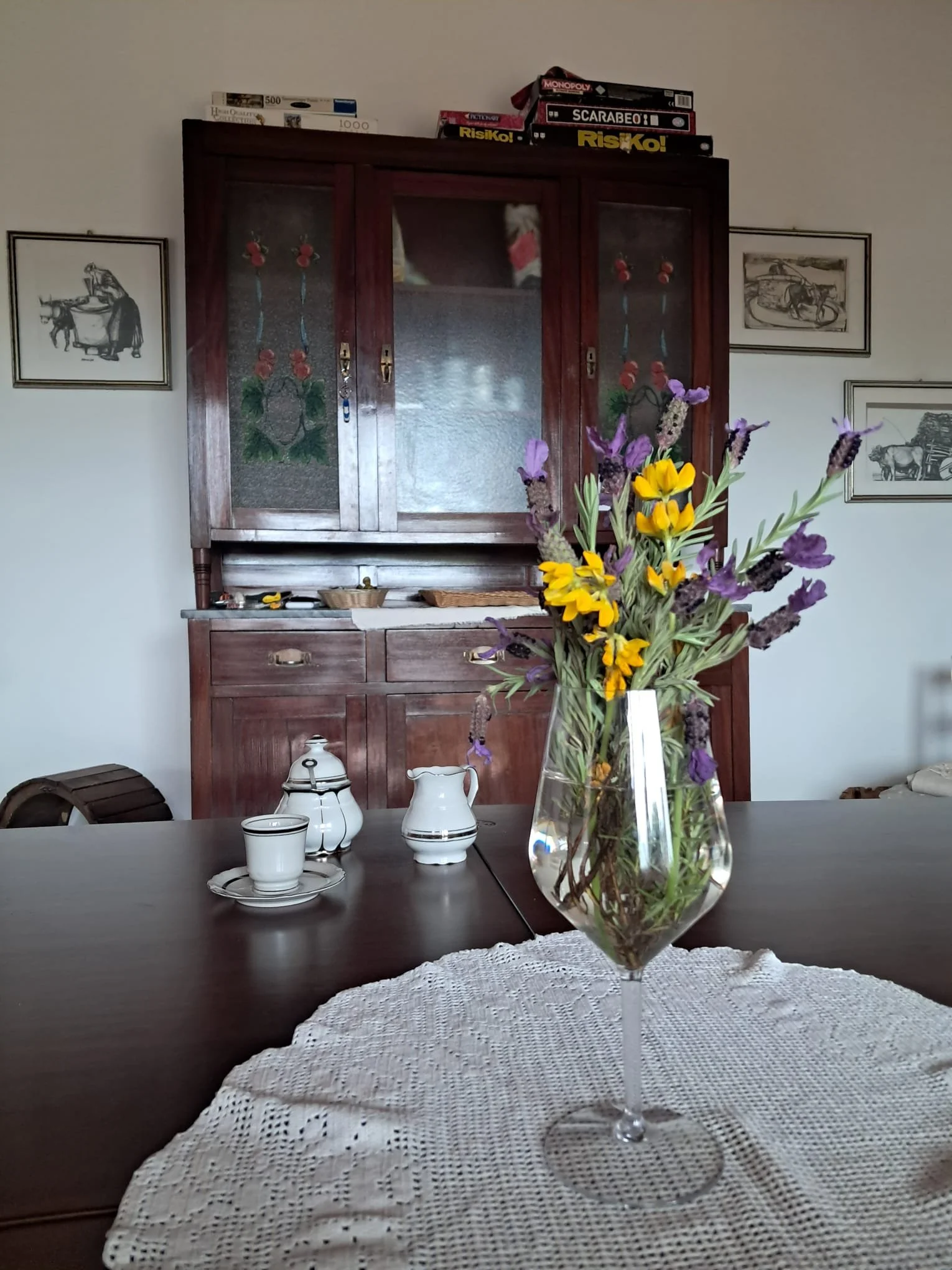Vaso con fiori freschi su tavolo in legno, con tazza, teiera e zuccheriera in porcellana bianca con dettagli neri, in una stanza con armadio in legno e fotografie alle pareti.