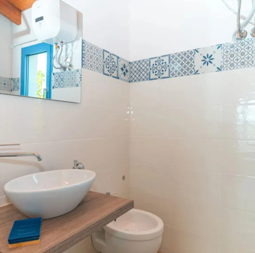 Bagno con lavabo a bacinella bianca sopra un piano in legno, specchio, water, pareti rivestite in piastrelle bianche e decorazioni blu.