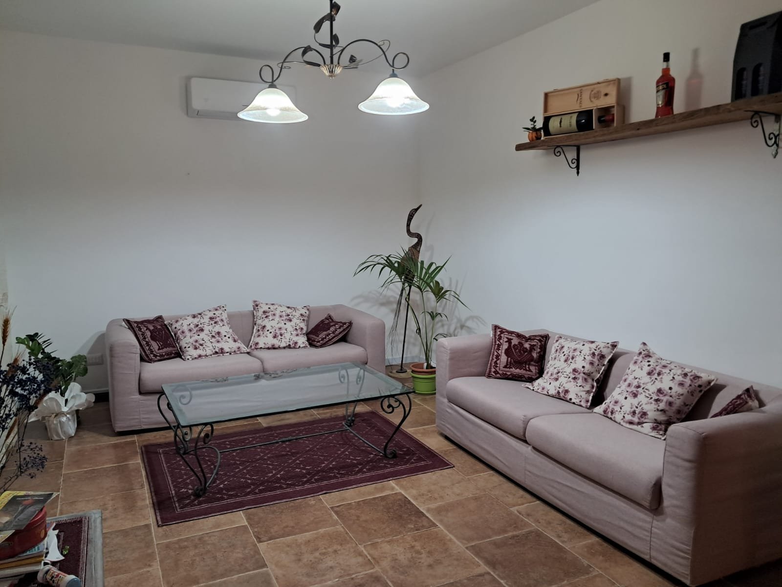 Soggiorno con due divani beige con cuscini decorativi, tavolino in vetro e ferro battuto, tappeto rosso e piante verdi. Illuminazione con lampadario a 3 luci. Esterno bianco e pareti con mensole in legno.