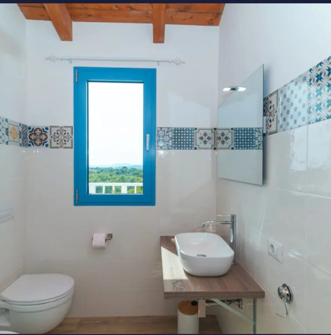 Bagno con finestra blu, lavabo su mobile in legno, water bianco, pareti con piastrelle decorate e un grande specchio.