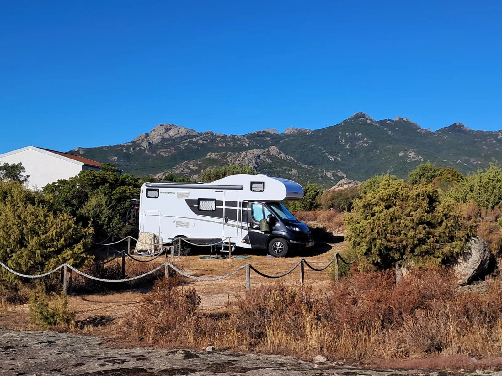 Camper bianco parcheggiato in un'area naturale con montagne sullo sfondo, cielo blu e vegetazione secca e arbusti