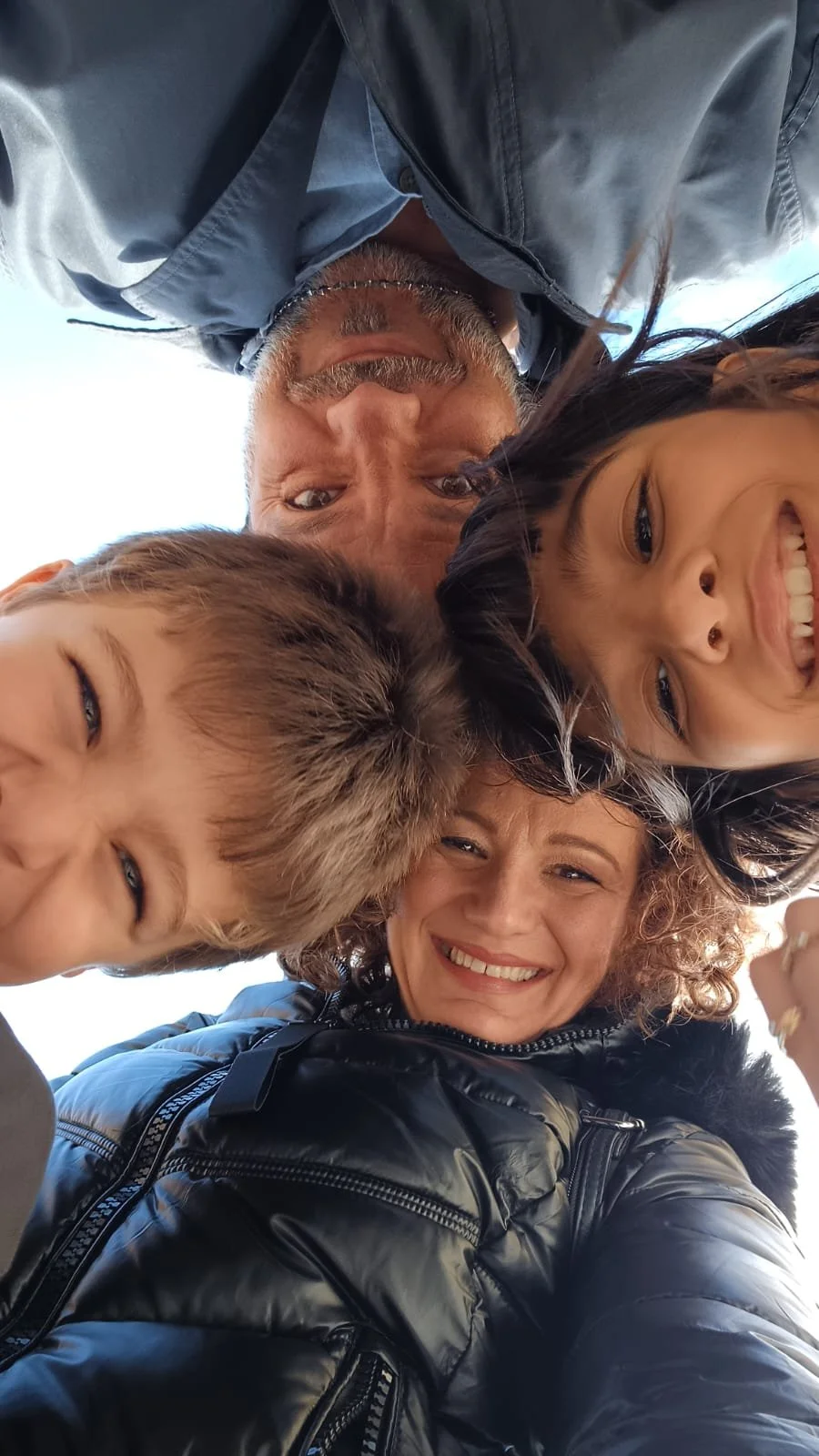 Una famiglia composta da due adulti e due bambini sorride e si riunisce in un abbraccio, con uno sfondo di cielo chiaro.