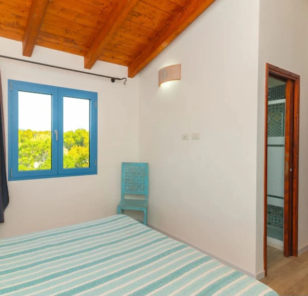 Camera da letto con pareti bianche, tetto in legno a vista, finestra con cornice blu che mostra una vista di alberi verdi, letto con copriletto azzurro e bianco, sedia azzurra in un angolo, porta che conduce a una stanza con piastrelle colorate.