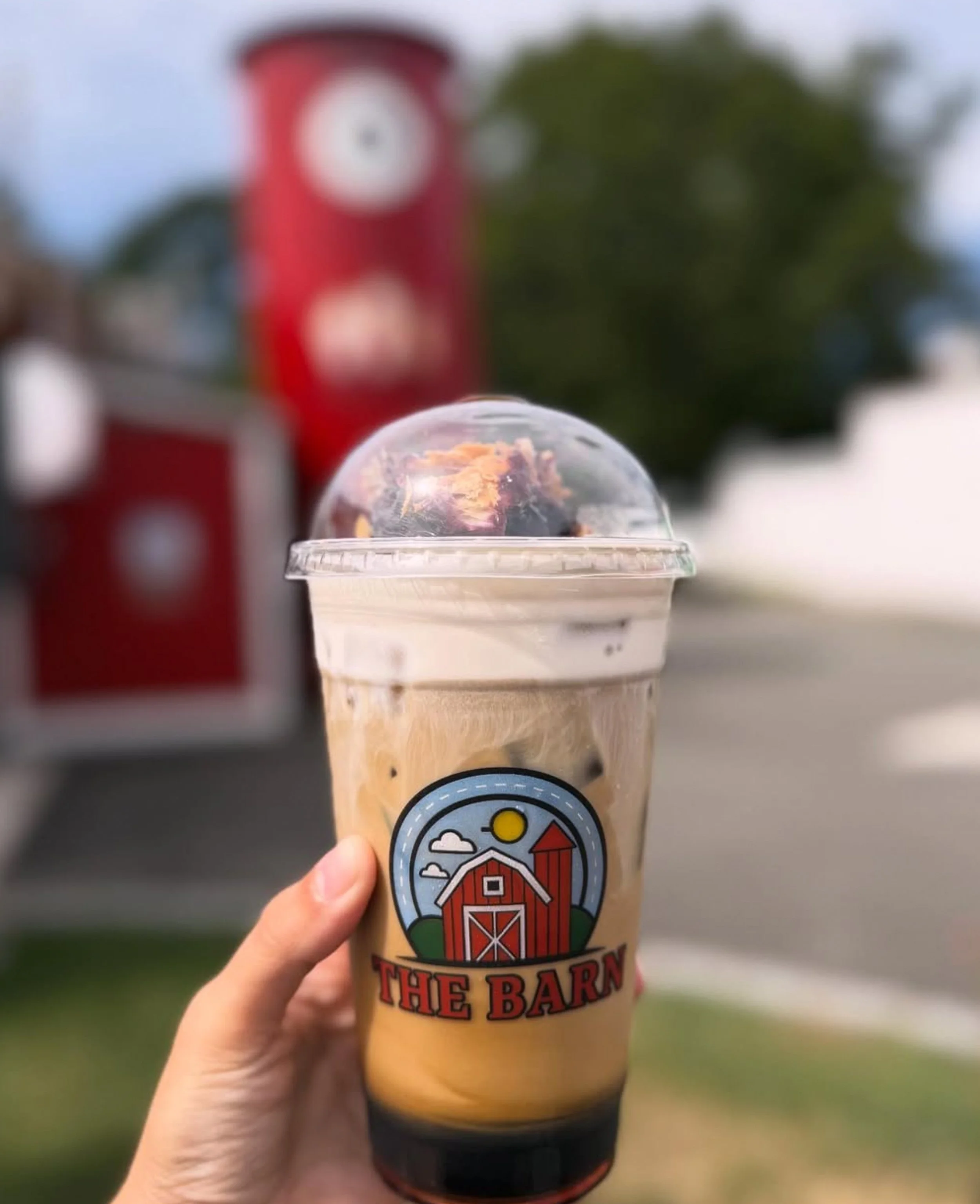 A hand holding a plastic cup of iced coffee with a logo that reads 'The Barn'. The background includes a red fire alarm box and a blurred outdoor scene with trees.