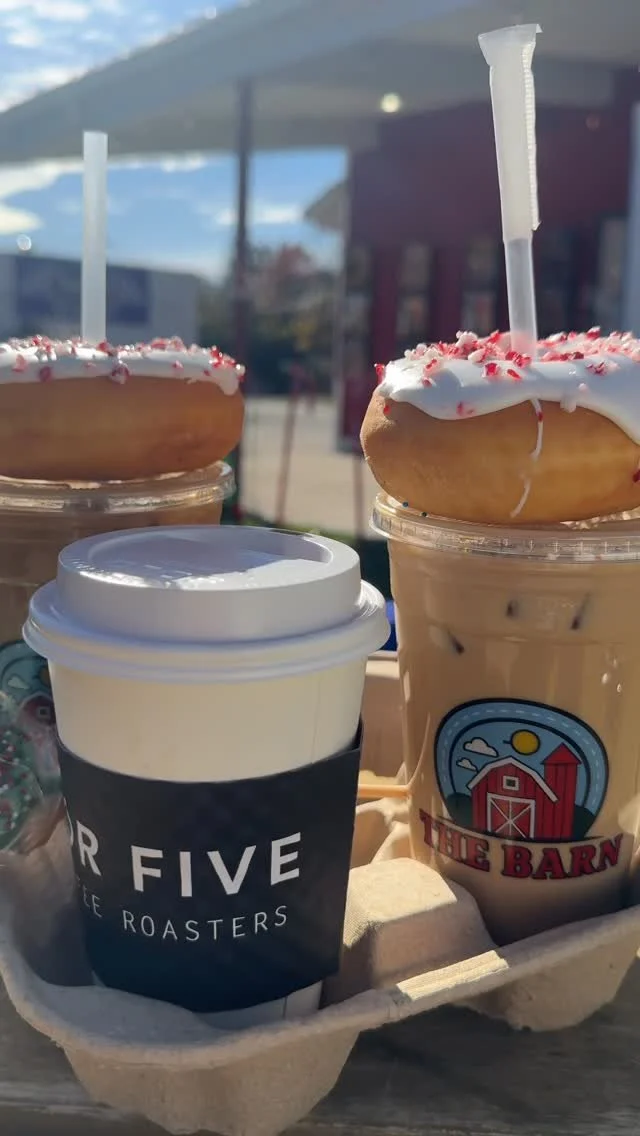 Two donuts with white icing and red sprinkles on top of iced coffee drinks with whipped cream, placed in a cardboard tray outdoors.