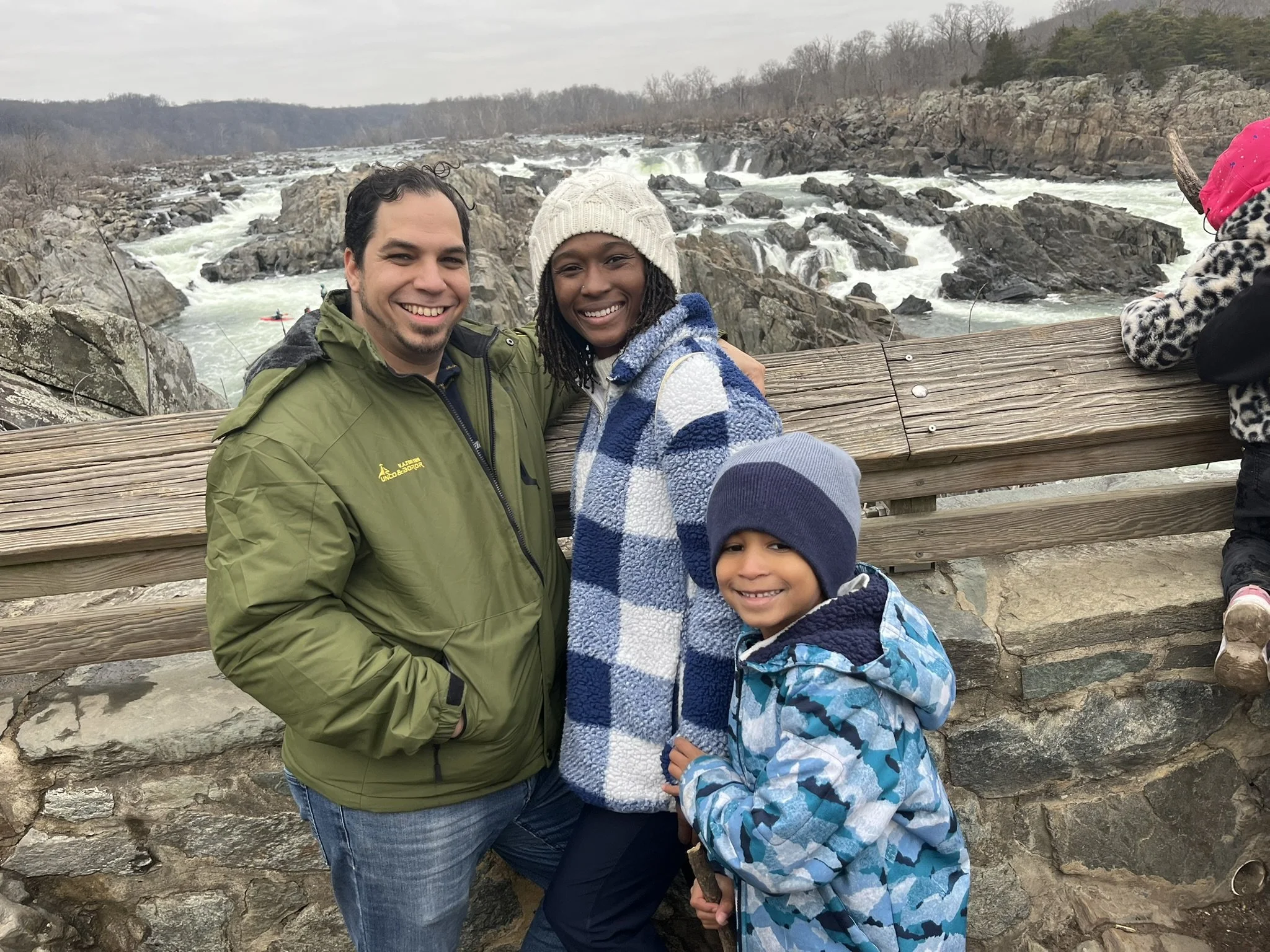 A family of three smiling on a bridge overlooking a rocky river with water rapids, wearing winter jackets and hats.