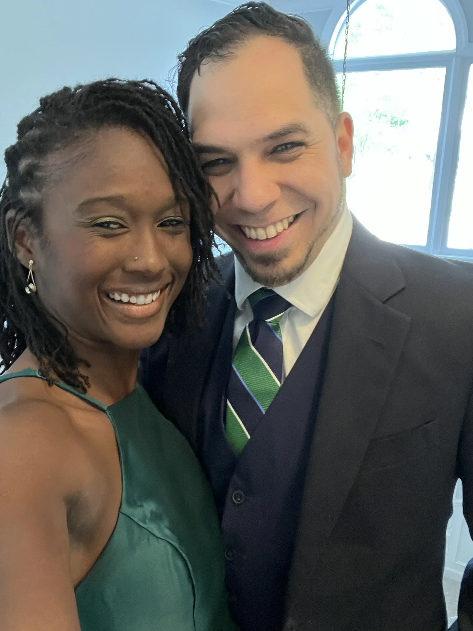 A smiling woman with dark skin, dreadlocked hair, and a nose piercing, in a green dress, standing close to a smiling man with light skin and short dark hair, wearing a suit and a striped tie, posing for a close-up selfie indoors near a window.