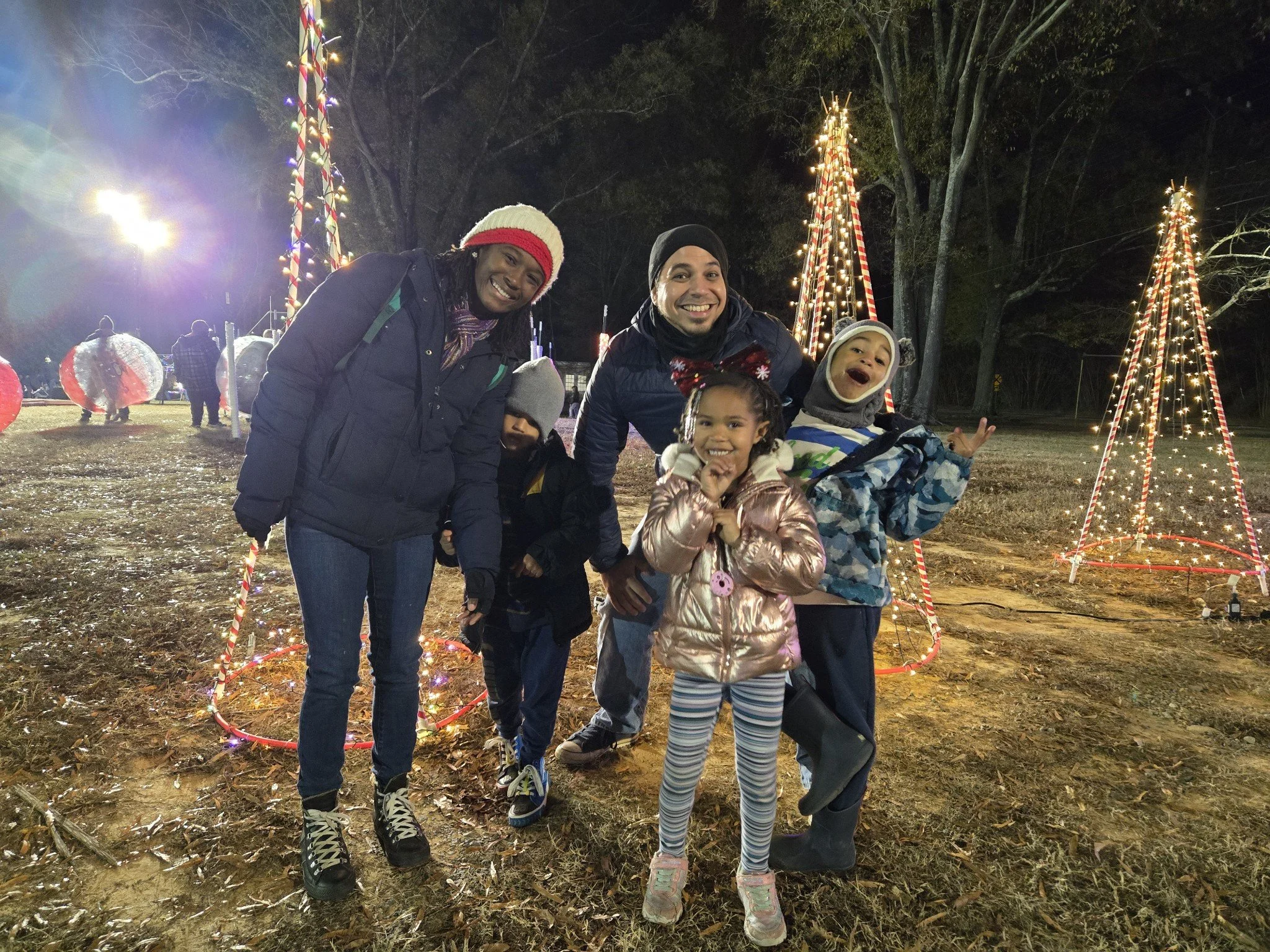 Family of five smiling and posing outdoors at night during a holiday event, with Christmas light displays and decorated Christmas trees in the background.
