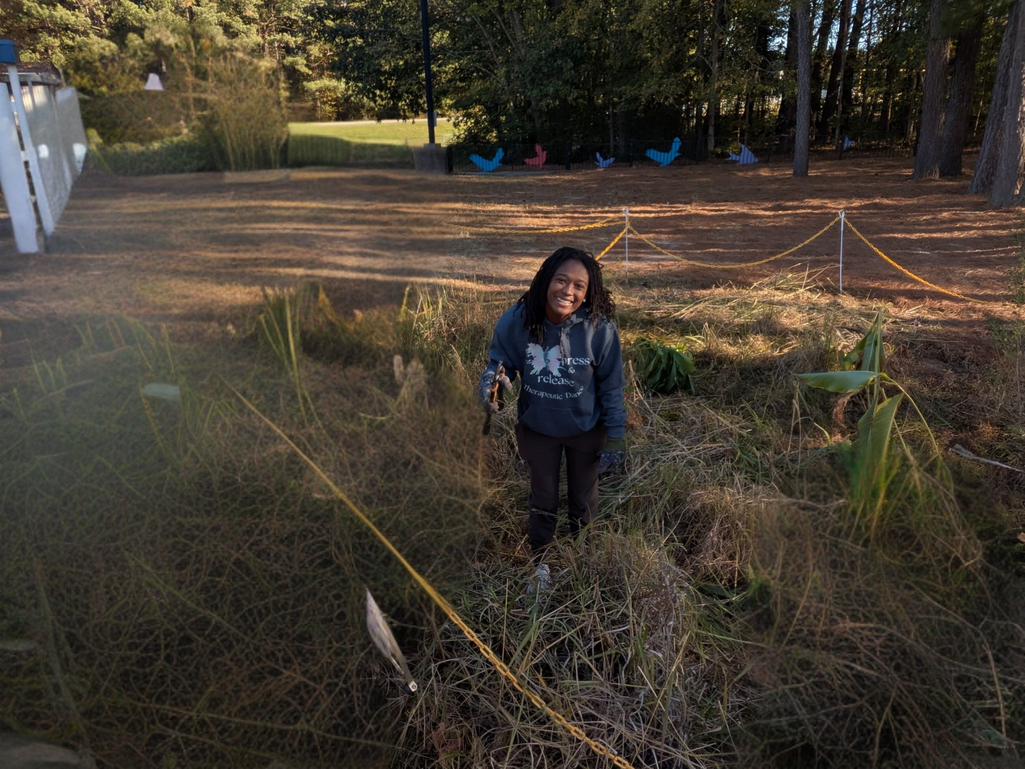 A woman with curly black hair wearing a blue sweatshirt and black pants smiling while holding a gardening tool, standing in a garden with tall grass and plants, behind a yellow rope barrier and trees.